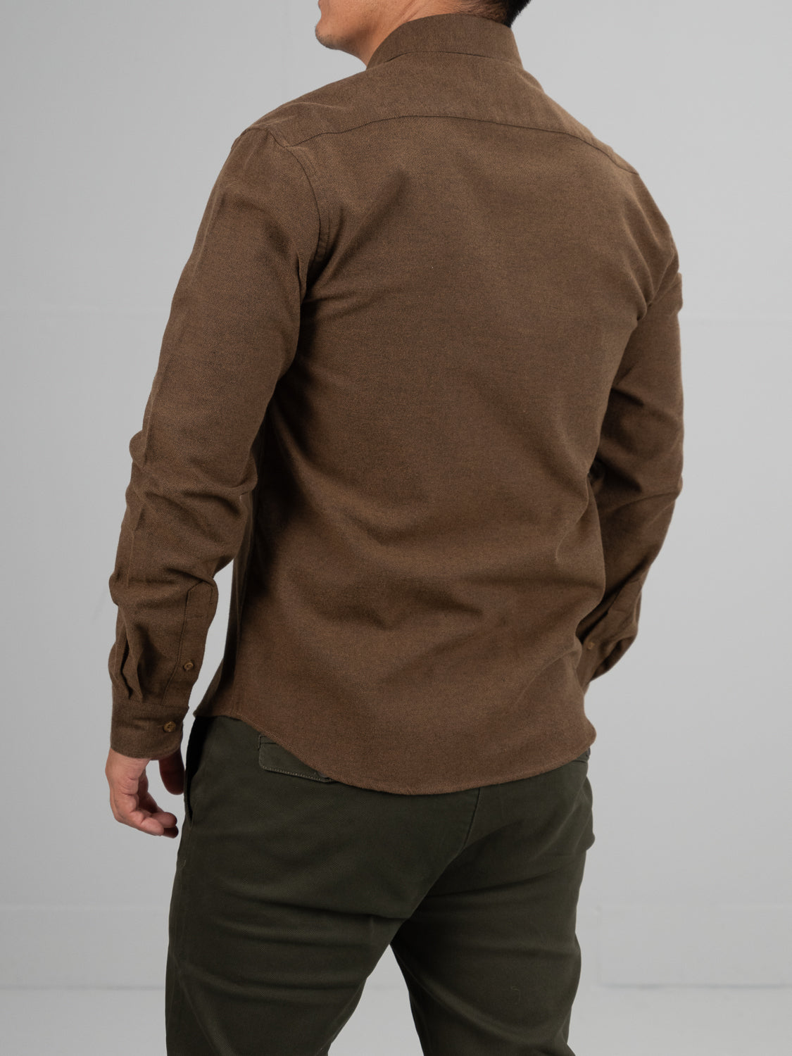 A person in an ID Menswear Magnus Brushed Twill Solid Flannel Shirt, brown with a button-down collar, and dark green pants stands against a light gray background, back to the camera.