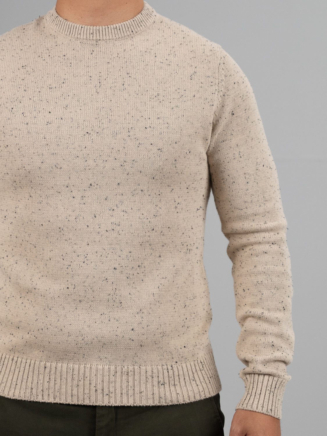 A person in a beige Malmo Speckled Cotton Sweater by ID Menswear with ribbed cuffs and hem and dark pants stands against a plain gray background; their head is not visible.