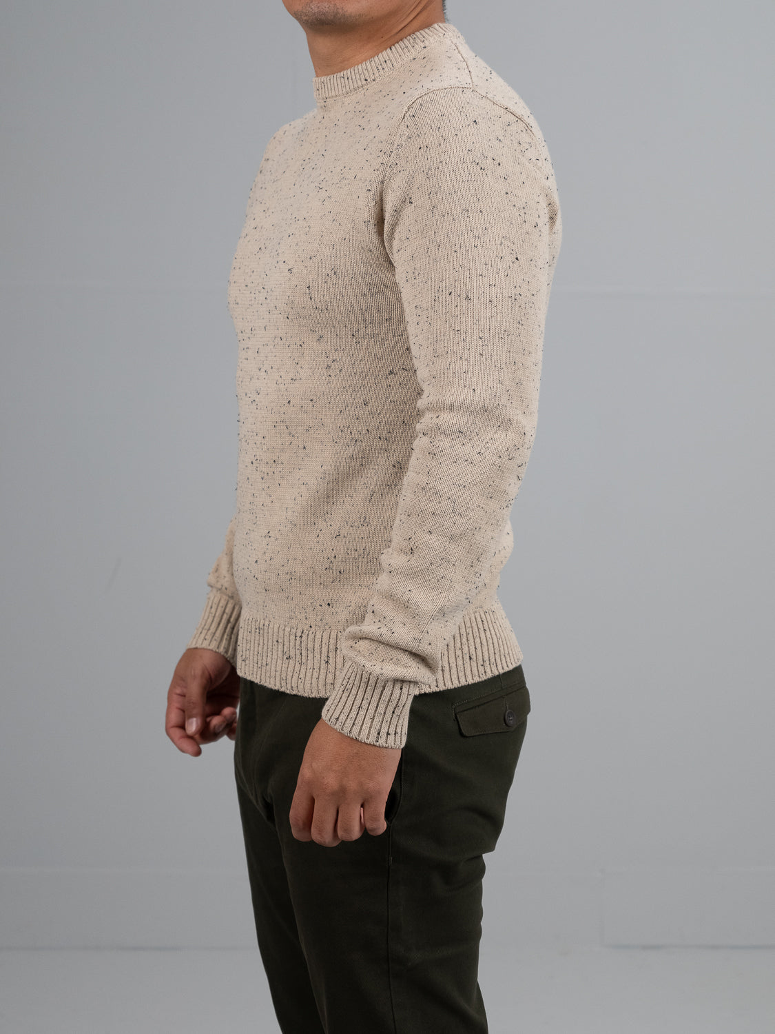 A person stands sideways against a plain gray background, wearing the ID Menswear Malmo Speckled Cotton Sweater in light beige and dark olive green pants with side pockets. Their face is partially out of the frame.