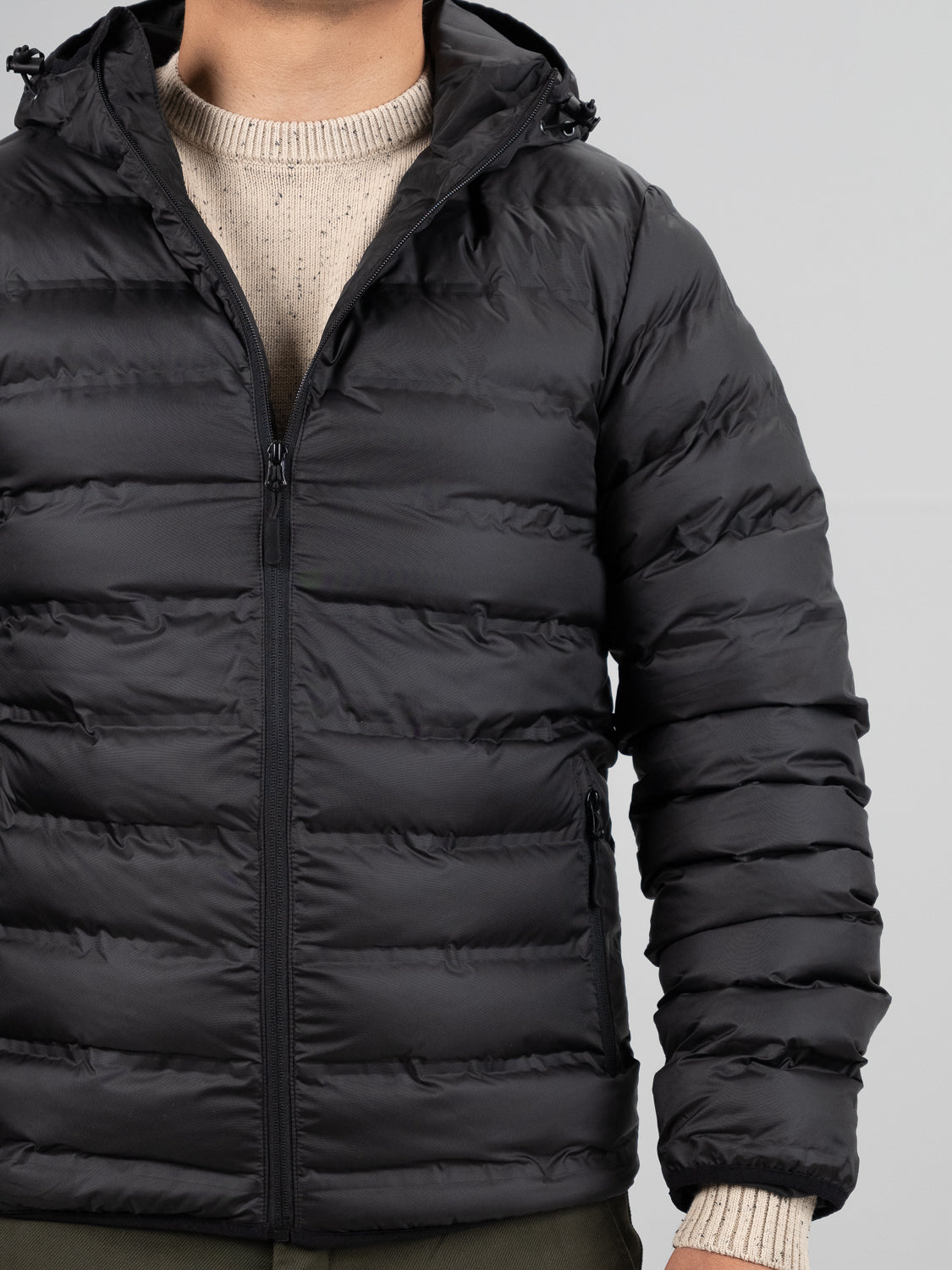 A person wears the Yukon Hooded Puffer Jacket by ID Menswear, a black lightweight regular fit puffer zipped over a beige knit sweater, shown shoulder to waist against a light background with their face not visible.