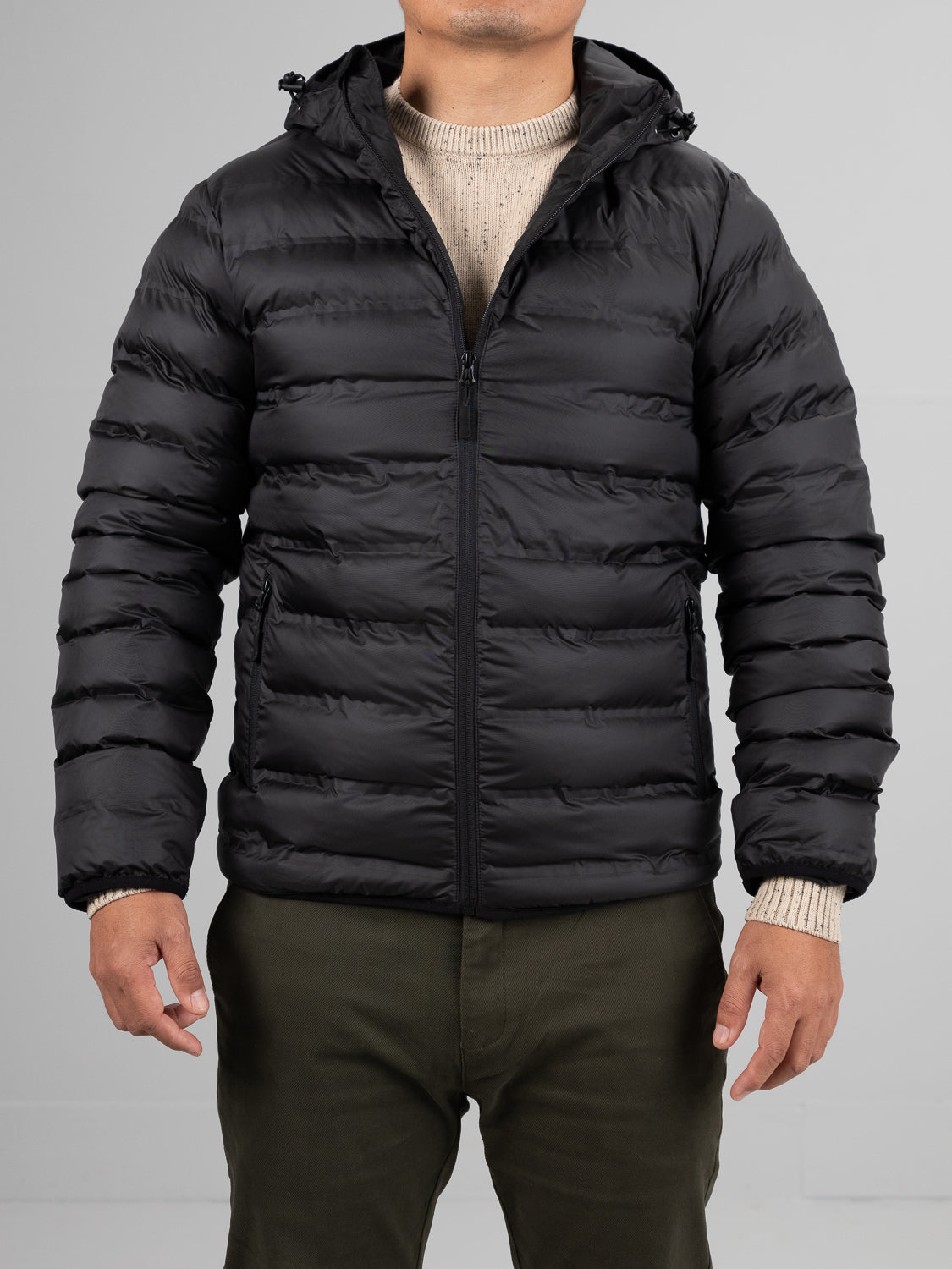 A person in the ID Menswear Yukon Hooded Puffer Jacket with a polyester shell stands facing forward over a beige sweater and olive green pants, arms relaxed. Regular fit and plain background highlight the outfit. Face not visible.