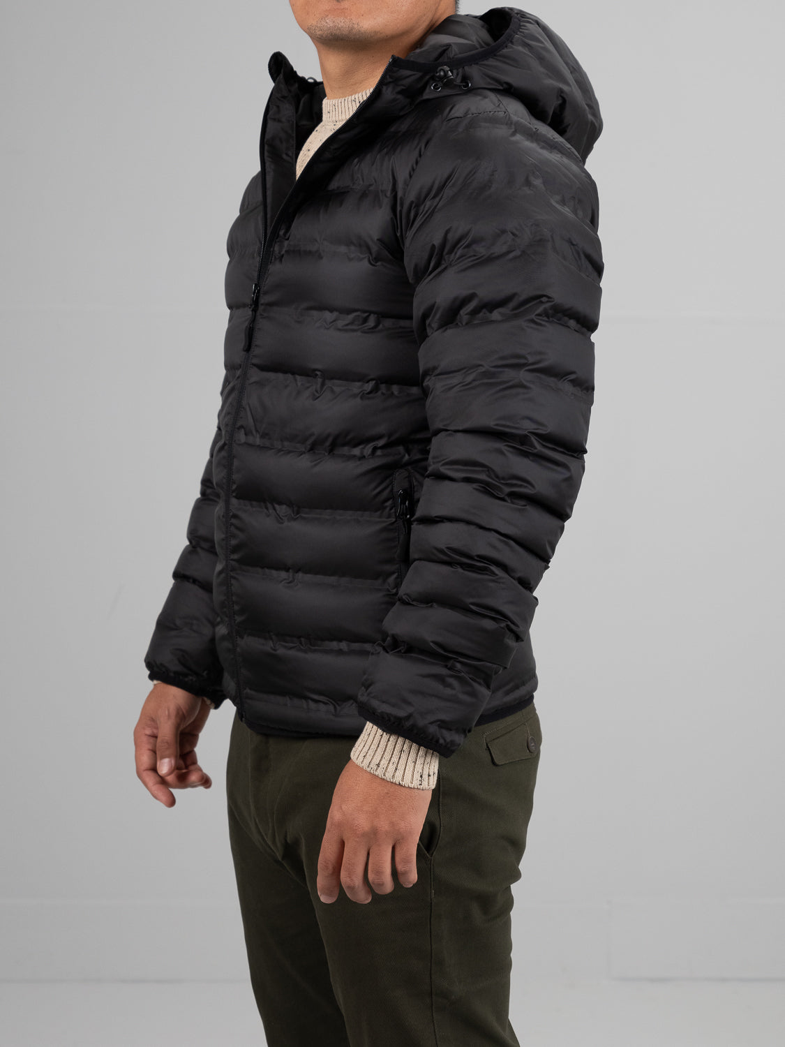 A person wears the Yukon Hooded Puffer Jacket by ID Menswear—a black, regular fit, hooded puffer with a polyester shell—over a beige knit sweater and olive green pants, standing against a plain light gray background.