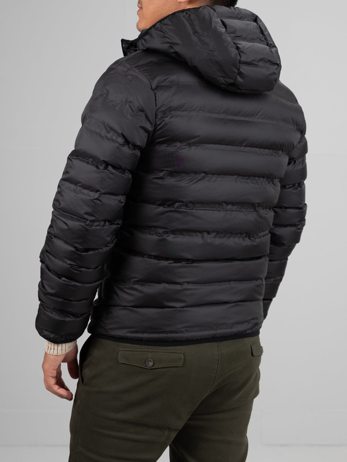 A person faces away, wearing the ID Menswear Yukon Hooded Puffer Jacket with a polyester shell and hood, paired with olive green pants against a plain light gray background.