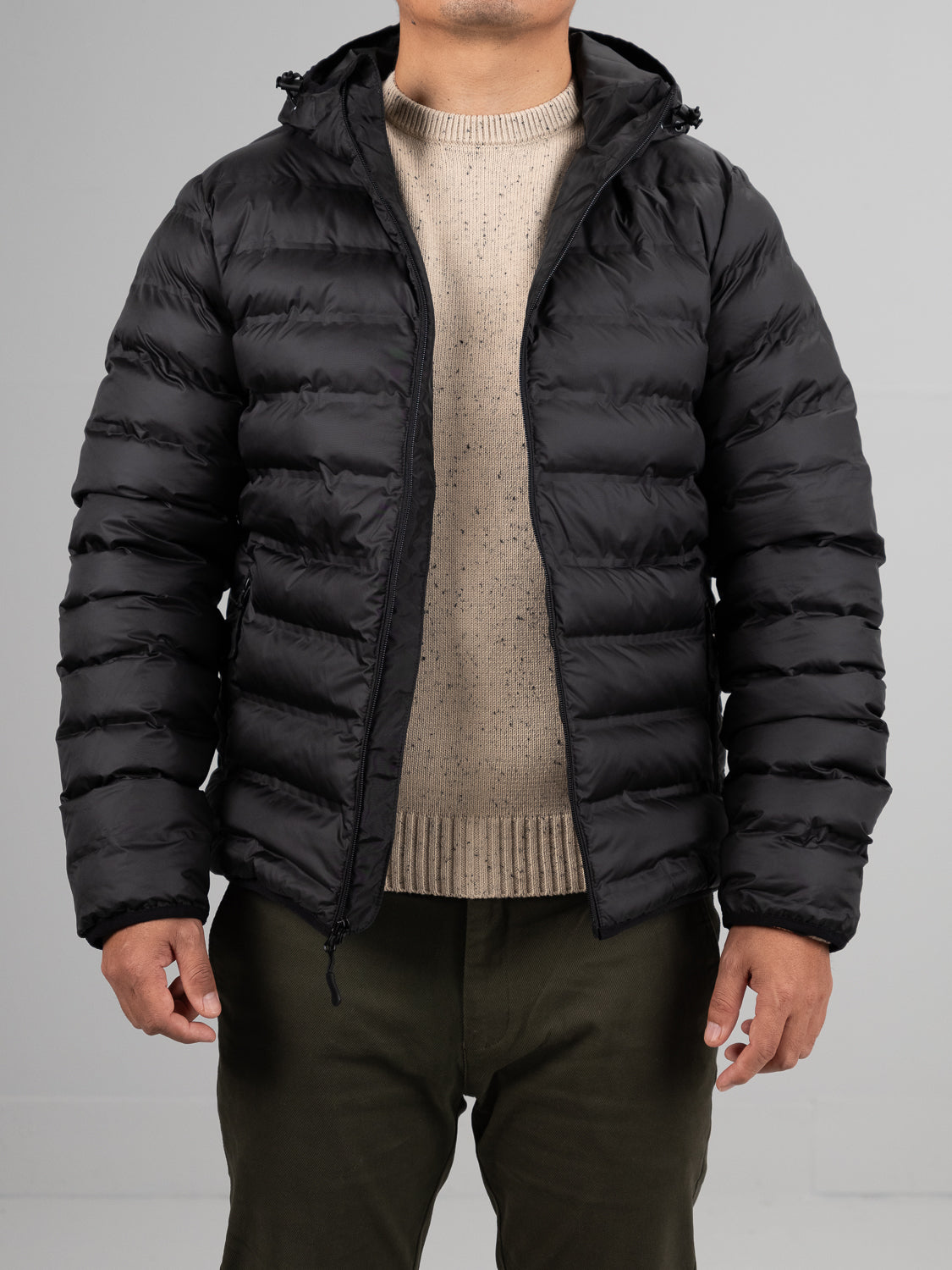 A person wearing the ID Menswear Yukon Hooded Puffer Jacket in black over a beige knit sweater and dark green pants stands facing forward against a plain light gray background, with their face not visible.