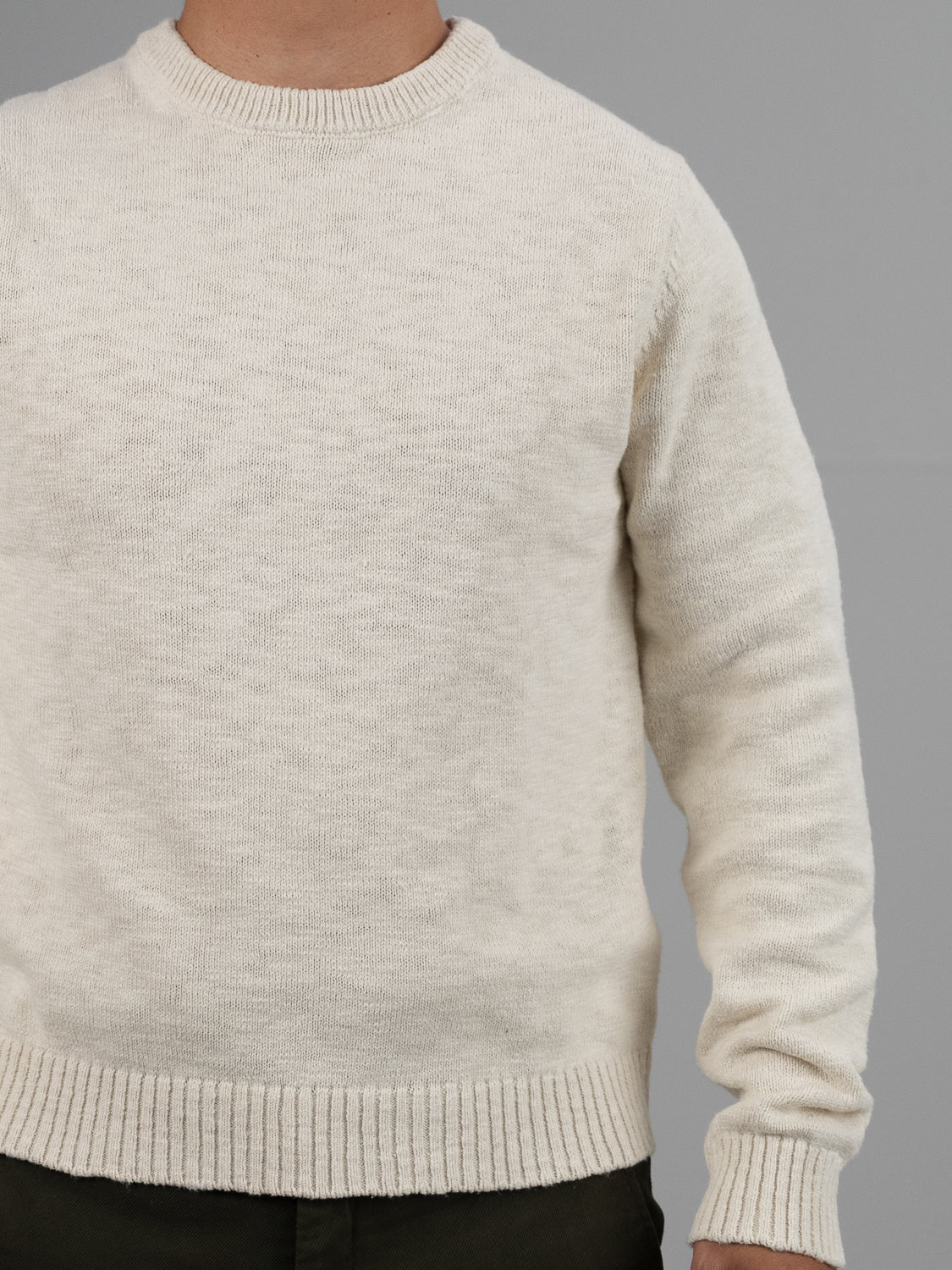 A person models the Odense Slub Yarn Sweater by ID Menswear, sporting a cream-colored, regular-fit crew neck. They pair it with dark pants against a gray backdrop, with their head out of the frame.