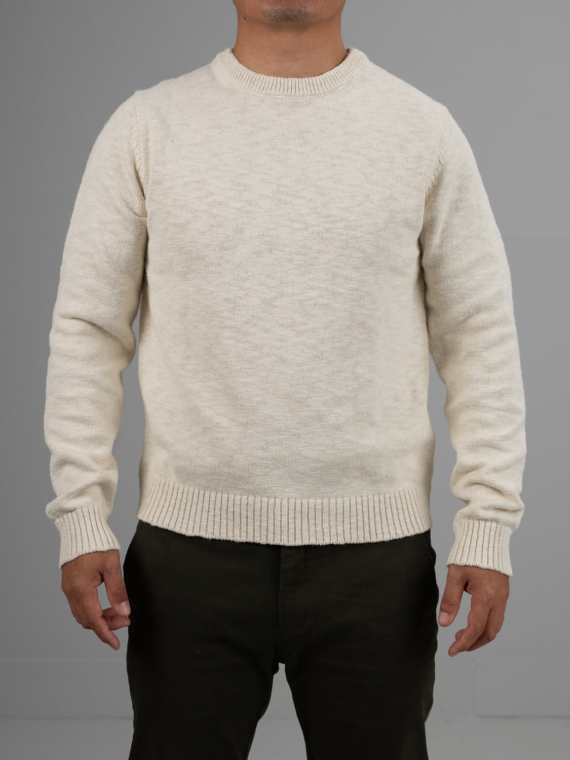 A person models the cream Odense Slub Yarn Sweater by ID Menswear against a neutral gray backdrop. It’s crafted from slub cotton with a ribbed crewneck, cuffs, and hem. Dark pants complement the regular-fit sweater, though their face is not visible.