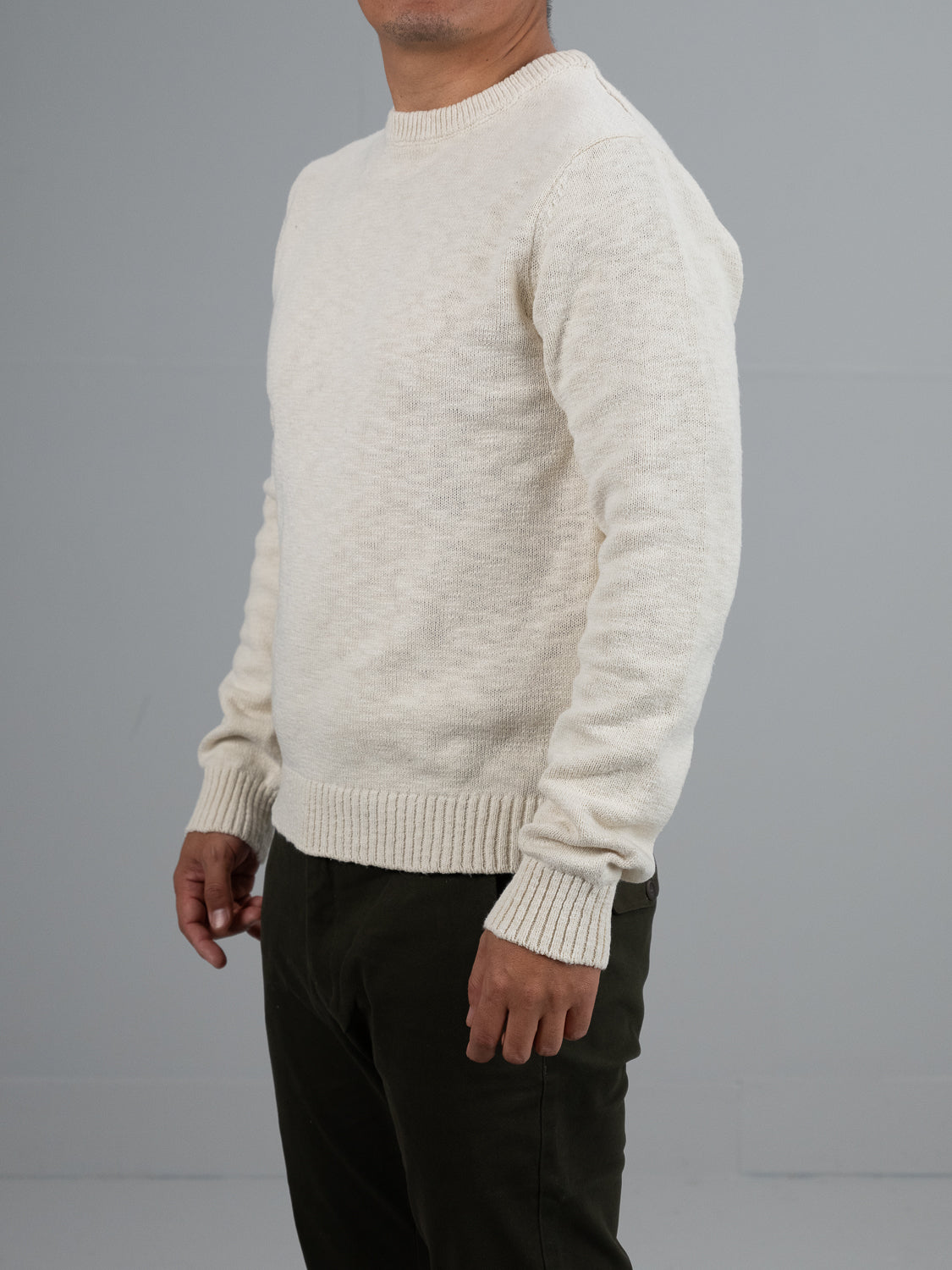 A person wears an ID Menswear Odense Slub Yarn Sweater, cream-colored with a crew neck and long sleeves, paired with dark pants against a neutral gray background. They stand slightly angled to the side with their face not visible.