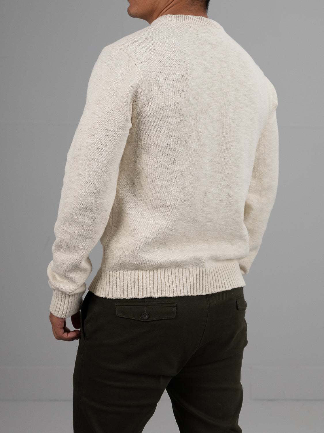 A person wearing an ID Menswear Odense Slub Yarn Sweater, in off-white with a ribbed hem and cuffs, alongside dark green pants stands facing away from the camera against a gray background.