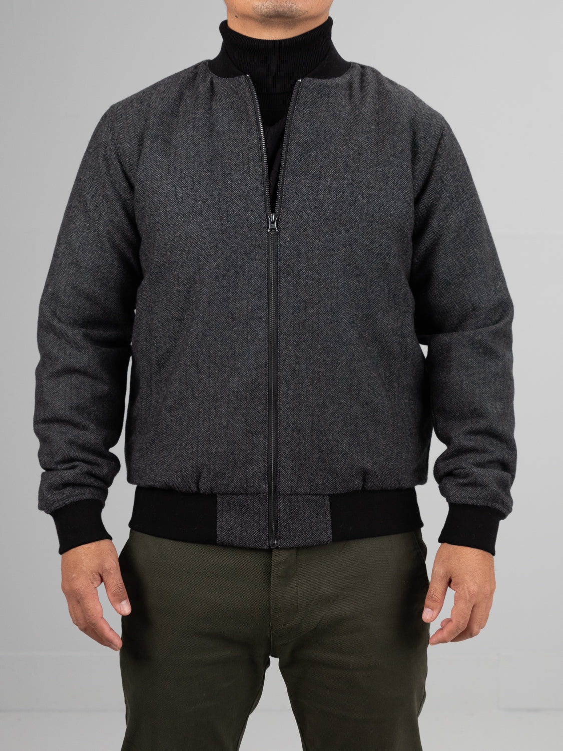 A person wearing an ID Menswear Montauk Wool Blend Jacket, featuring a wool blend tweed with black zipper and cuffs, stands against a neutral background. The jacket is stylishly paired with dark green pants and a black turtleneck, the hidden face highlighting the ensembles lightweight comfort.