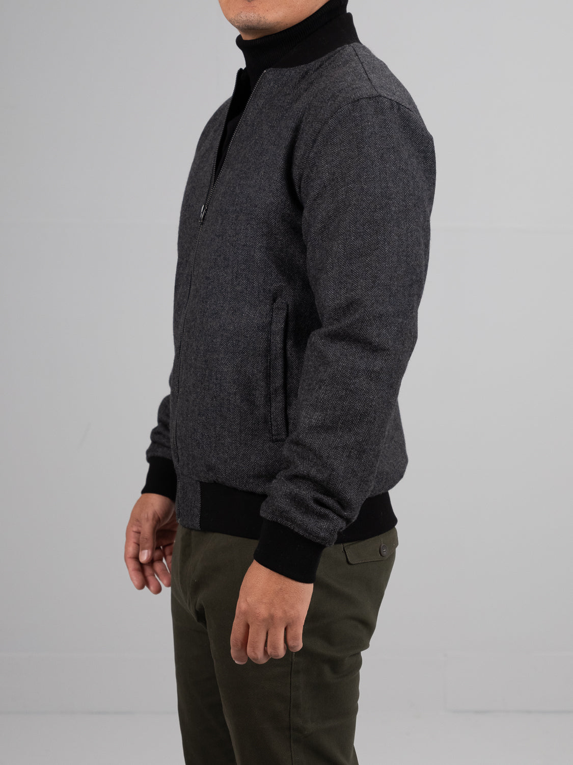 A person in an ID Menswear Montauk Wool Blend Jacket over a black turtleneck stands against a plain backdrop. This dark gray zip-up jacket features ribbed cuffs, offering lightweight comfort and style, paired with olive green pants.