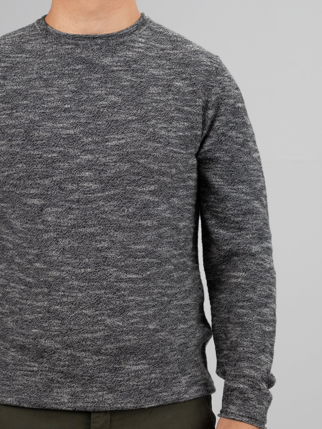 A person is wearing the Maverick Rolled Neck Sweater by ID Menswear, made from gray textured Merino wool, with lightweight warmth, and olive green pants. They stand against a plain light gray background, with their face not visible.