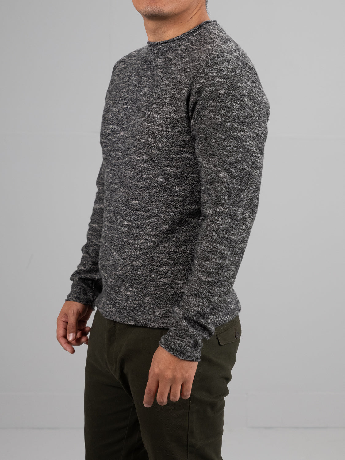 A person in an ID Menswear Maverick Rolled Neck Sweater and dark green pants stands against a gray backdrop. The sweaters lightweight warmth adds comfort as they pose slightly sideways, their left arm relaxed by their side.