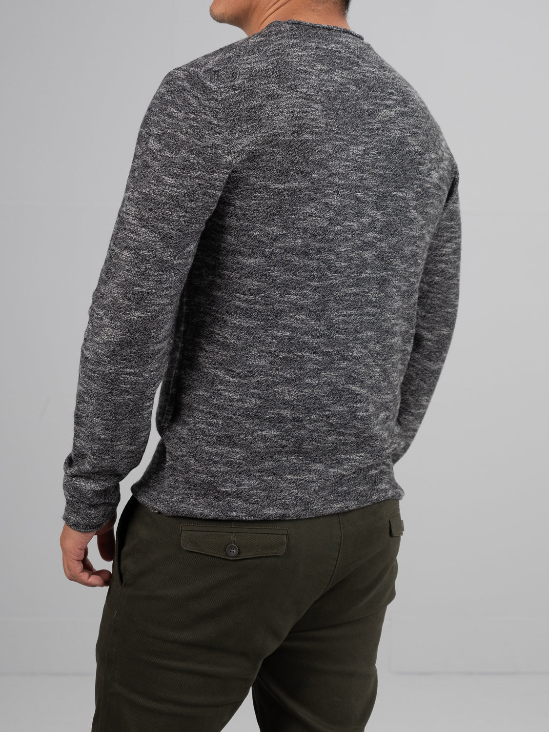 A person with short black hair stands facing away, draped in ID Menswear’s Maverick Rolled Neck Sweater, crafted from Merino wool for lightweight warmth. Paired with dark green pants, the ensemble blends seamlessly into the plain light gray background.