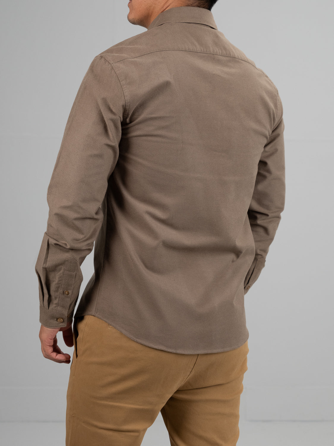 A person wearing a Milo Brushed Oxford Flannel Cotton Shirt by ID Menswear and tan pants faces away. The brown shirt features long sleeves with buttoned cuffs, perfect for any occasion. A plain gray background complements the attire.