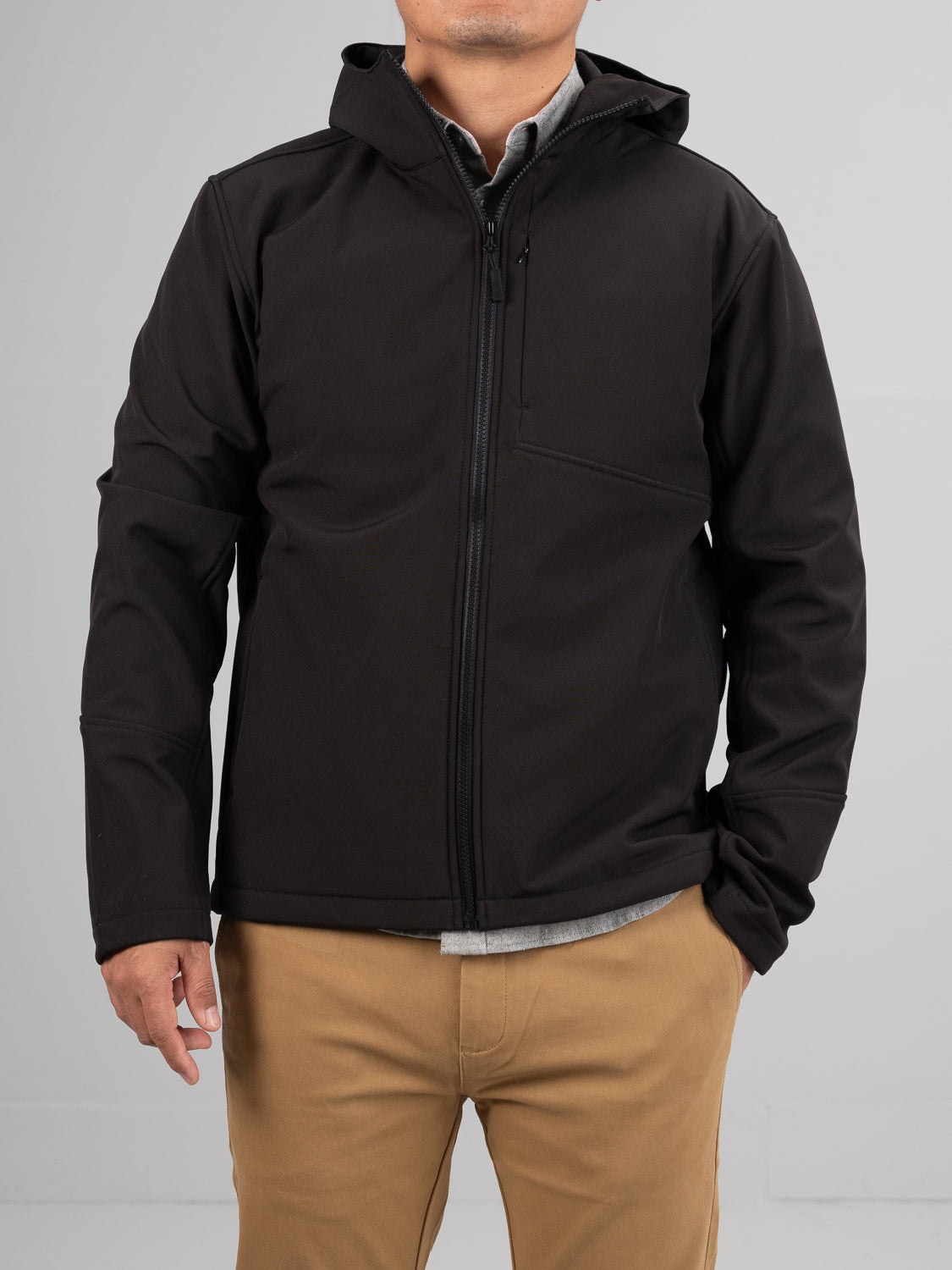 A person in a black Fairbanks Soft Shell Hooded Jacket by ID Menswear and khaki pants stands against a plain gray background. The jackets sleek, minimalist design features a regular fit and an extra chest pocket. Their hands rest relaxed by their sides.
