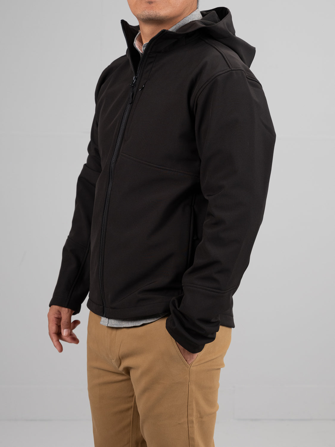 A person in an ID Menswear Fairbanks Soft Shell Hooded Jacket in black and beige pants stands against a plain background. The jacket, with its regular fit, features a front zipper, long sleeves, and an extra chest pocket. Their head is slightly turned, with hands relaxed at their sides.