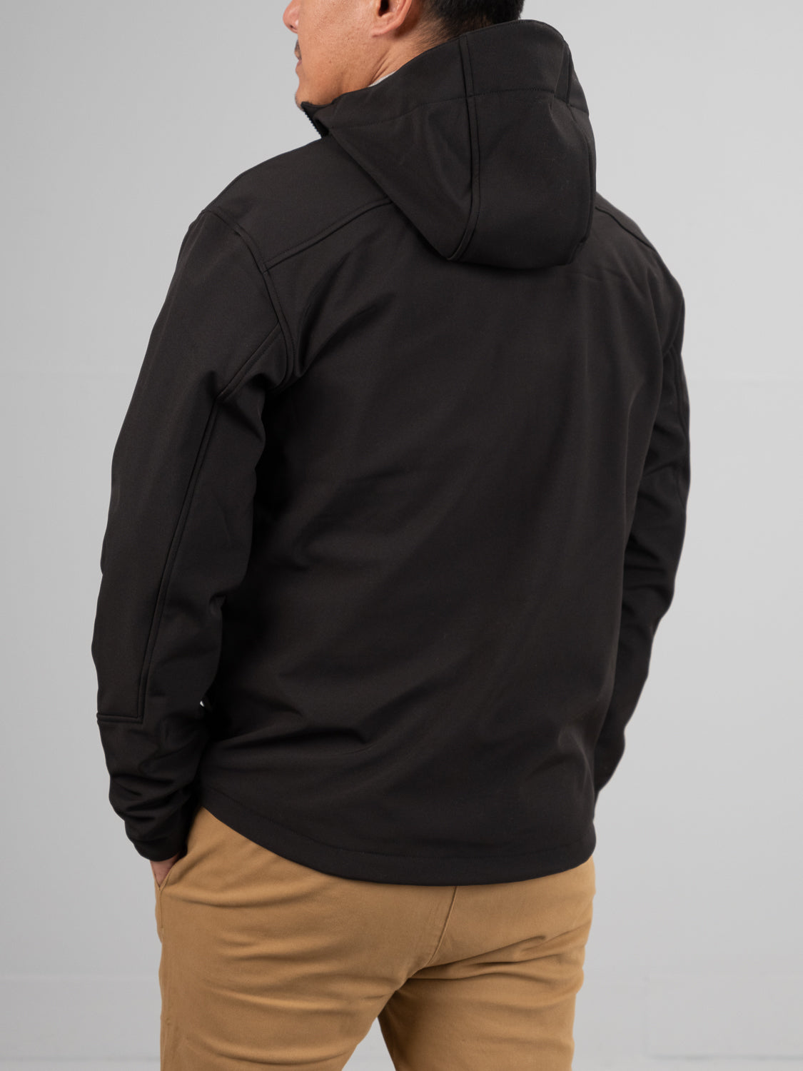 A person in ID Menswears Fairbanks Soft Shell Hooded Jacket, dark black color, and khaki pants stands with their back to us. The jackets regular fit provides comfort and style against the plain, light-colored background.