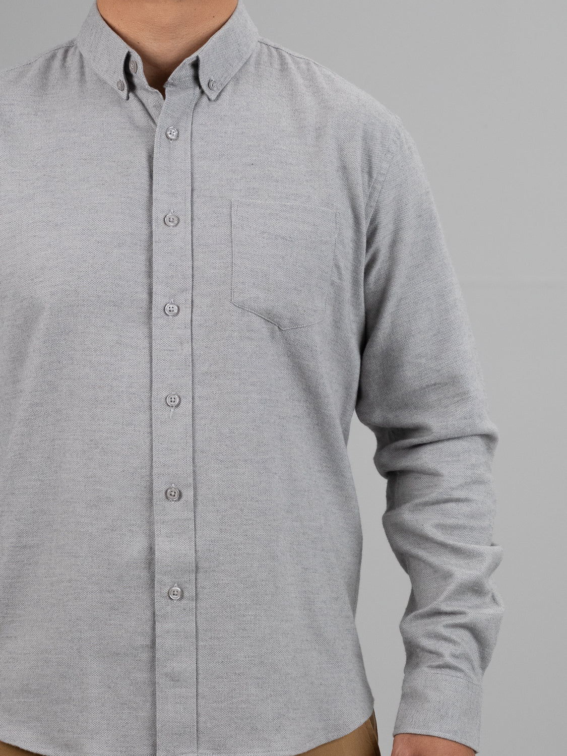 An individual stands against a gray backdrop wearing the Amir Solid Flannel Textured Dobby Shirt by ID Menswear. The light gray button-up, known for its lightweight comfort and featuring a left-side pocket, is tucked into tan pants. Only the upper body is visible.
