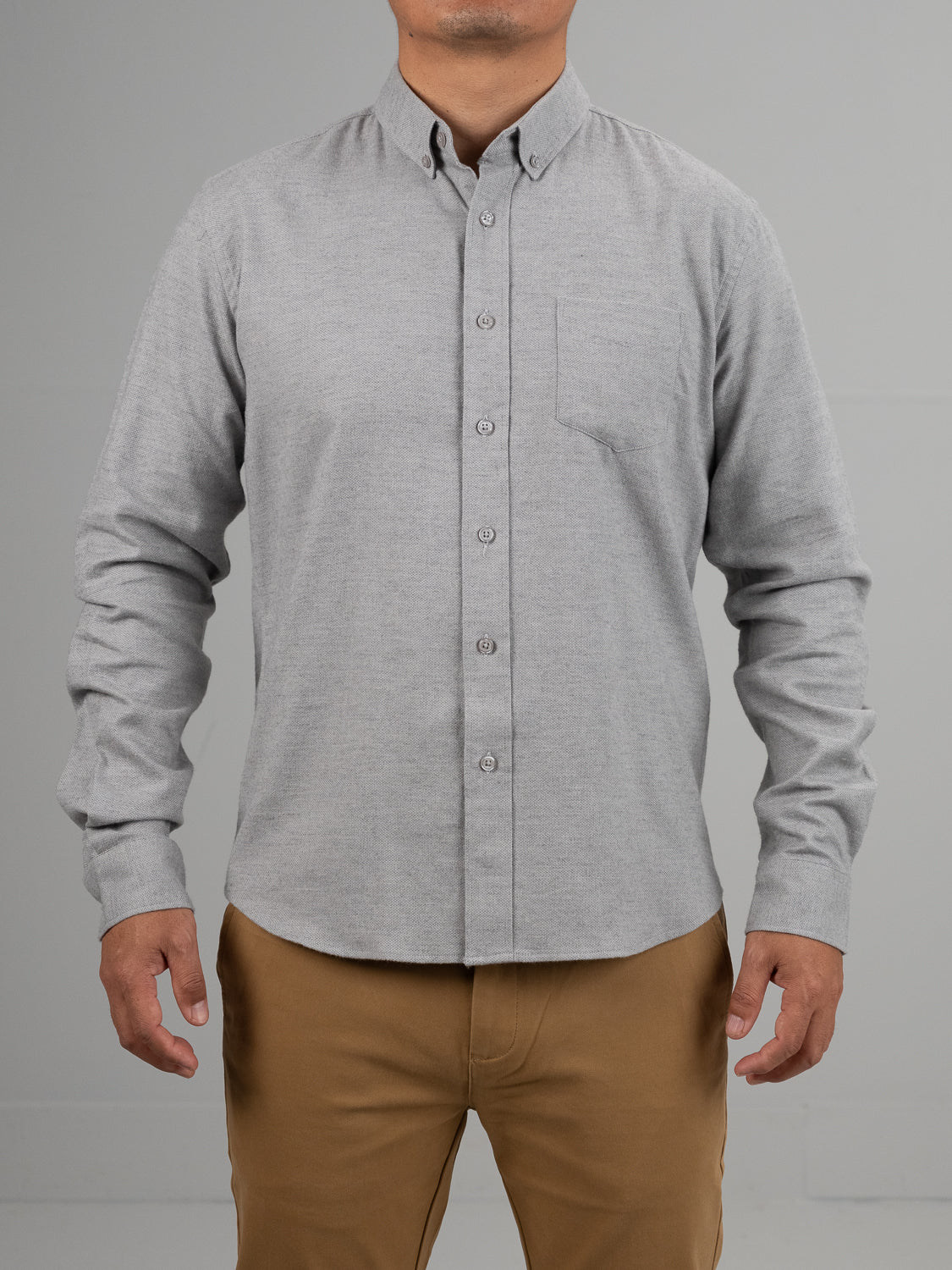 A person stands against a plain backdrop in the ID Menswear Amir Solid Flannel Textured Dobby Shirt, featuring long sleeves and a chest pocket. This light gray button-up top is paired with light brown pants, and their arms rest relaxed at their sides.
