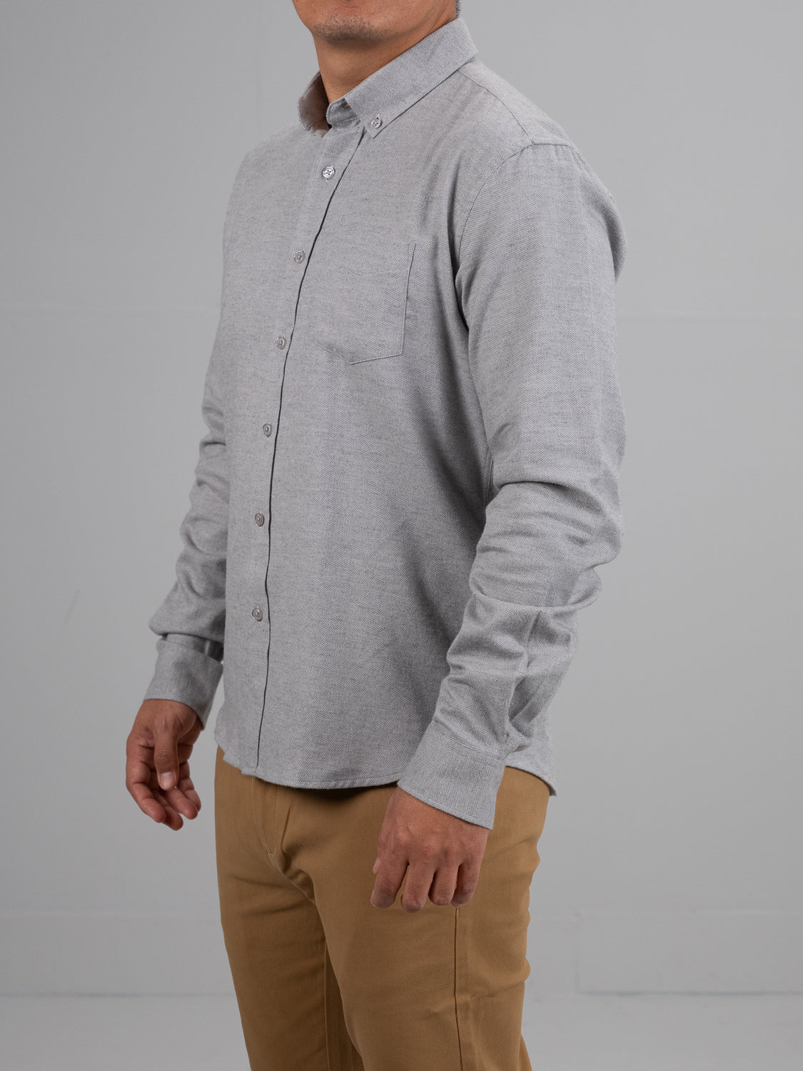 A person in an Amir Solid Flannel Textured Dobby Shirt from ID Menswear, light gray with a chest pocket, and beige pants stands against a plain background. The neutral-tone casual outfit offers lightweight comfort as the individual faces slightly left.