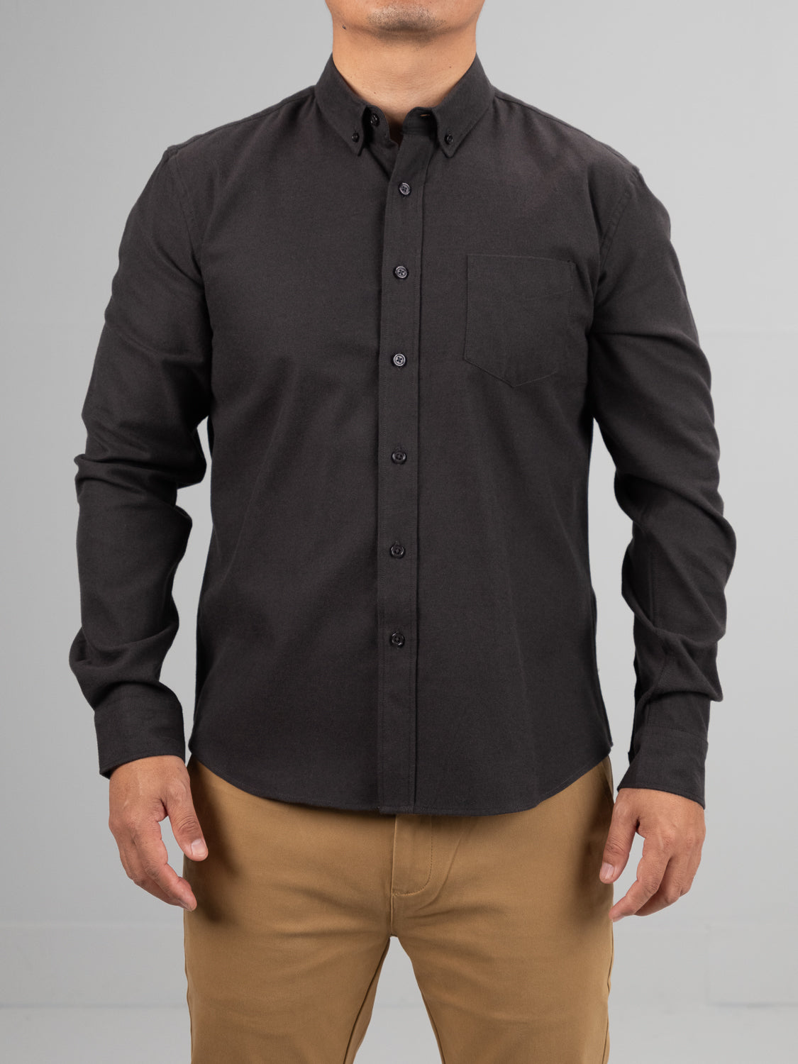 A person is wearing the Magnus Brushed Twill Solid Flannel Shirt by ID Menswear in black, featuring long sleeves and a chest pocket. The shirt is paired with tan pants against a plain, light gray background, with the persons head not visible.