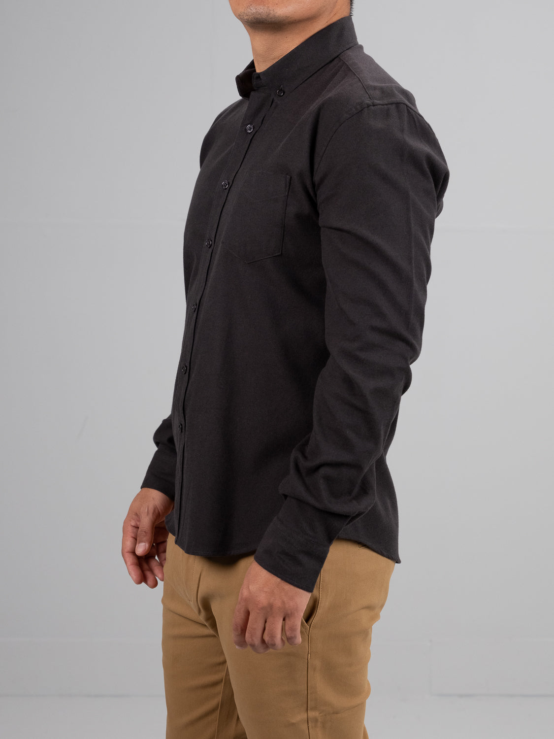 In a neutral-colored room, someone wears ID Menswears Magnus Brushed Twill Solid Flannel Shirt in black, featuring a button-down collar, paired with khaki pants. They are slightly turned to the left, keeping their face out of view.