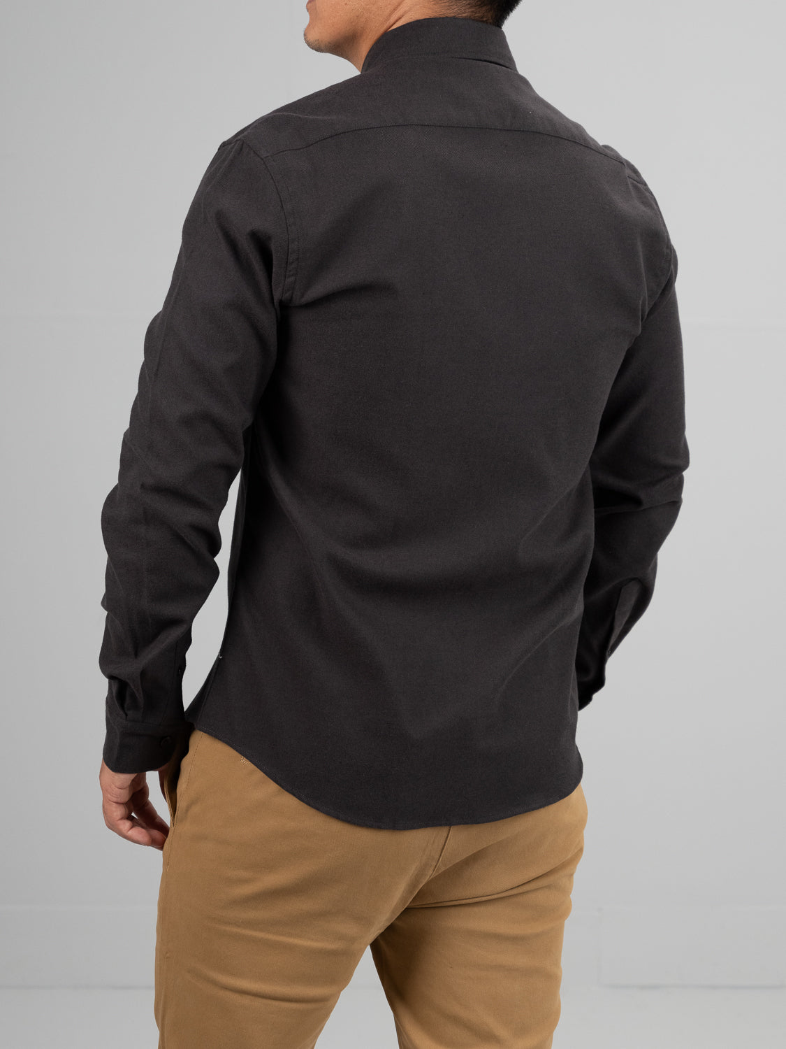 A person in a Magnus Brushed Twill Solid Flannel Shirt by ID Menswear, featuring long sleeves and a button-down collar, stands with their back to the camera. The dark shirt is well-fitted and smooth against a plain gray background, paired with beige pants.