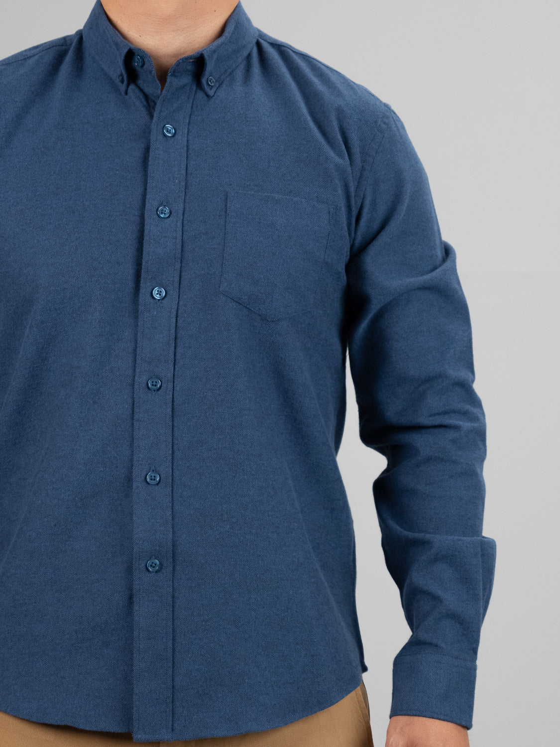A person is wearing a Magnus Brushed Twill Solid Flannel Shirt by ID Menswear, a buttoned-up, long-sleeve blue shirt with a stiff button-down collar and front pocket, paired with khaki pants against a plain gray backdrop.