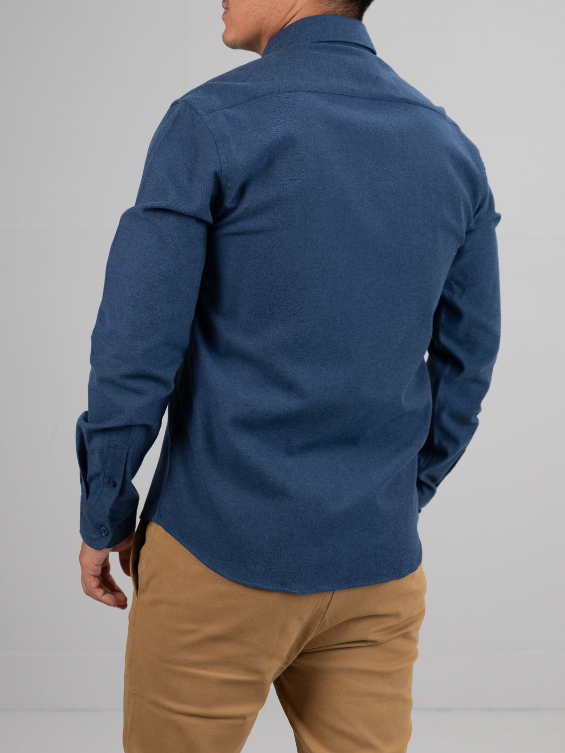 A person is shown from behind wearing the Magnus Brushed Twill Solid Flannel Shirt by ID Menswear, paired with tan pants. The blue shirt features a classic button-down collar and buttoned cuffs against a plain gray background.