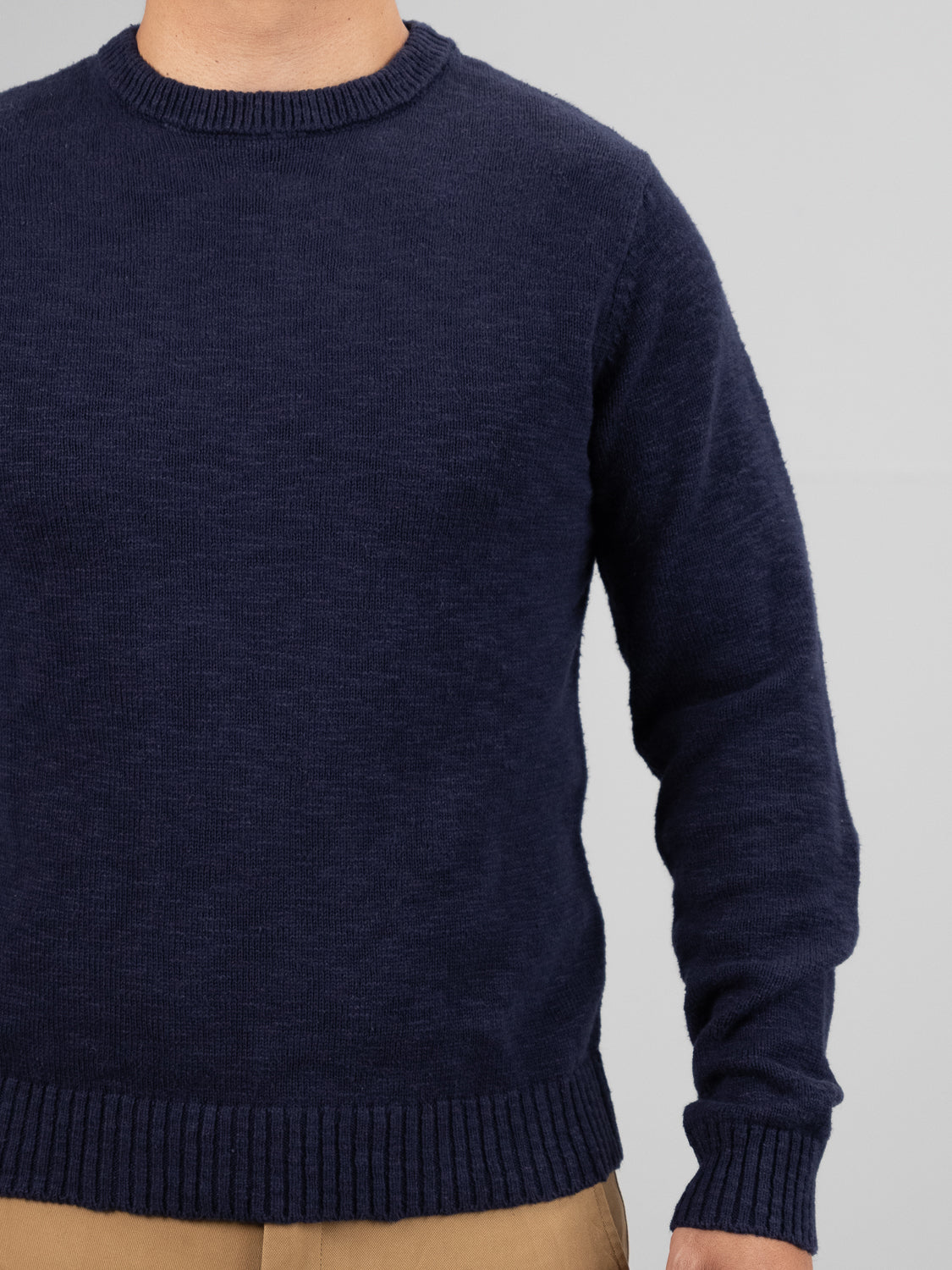 A person is shown from the shoulders down against a gray background wearing ID Menswears Odense Slub Yarn Sweater in dark blue with beige pants.