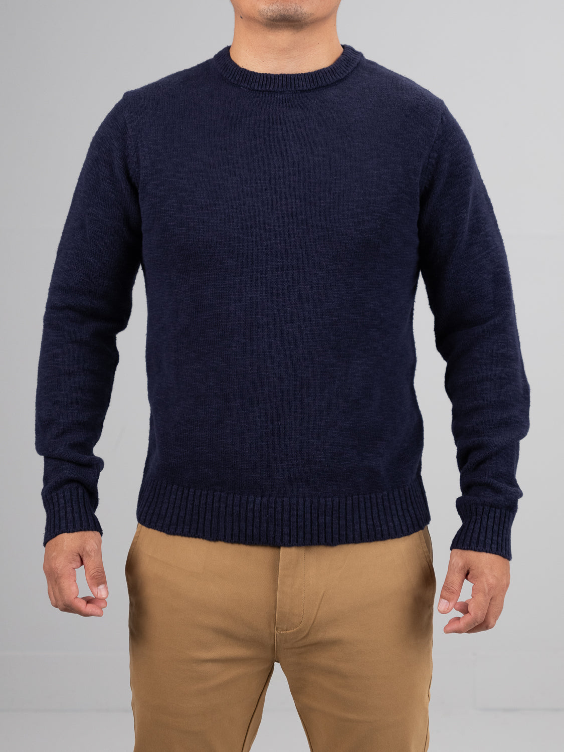 A person in a navy blue Odense Slub Yarn Sweater by ID Menswear, featuring ribbed cuffs, along with khaki pants, stands against a gray backdrop. The sweater is regular fit, crew neck, and made from soft slub cotton. The persons head is not visible in the image.