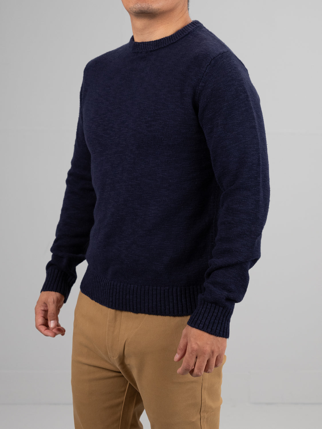A person wears the Odense Slub Yarn Sweater by ID Menswear, a dark blue, regular fit crew neck, paired with beige pants. The image captures their upper body and arms against a neutral gray background.