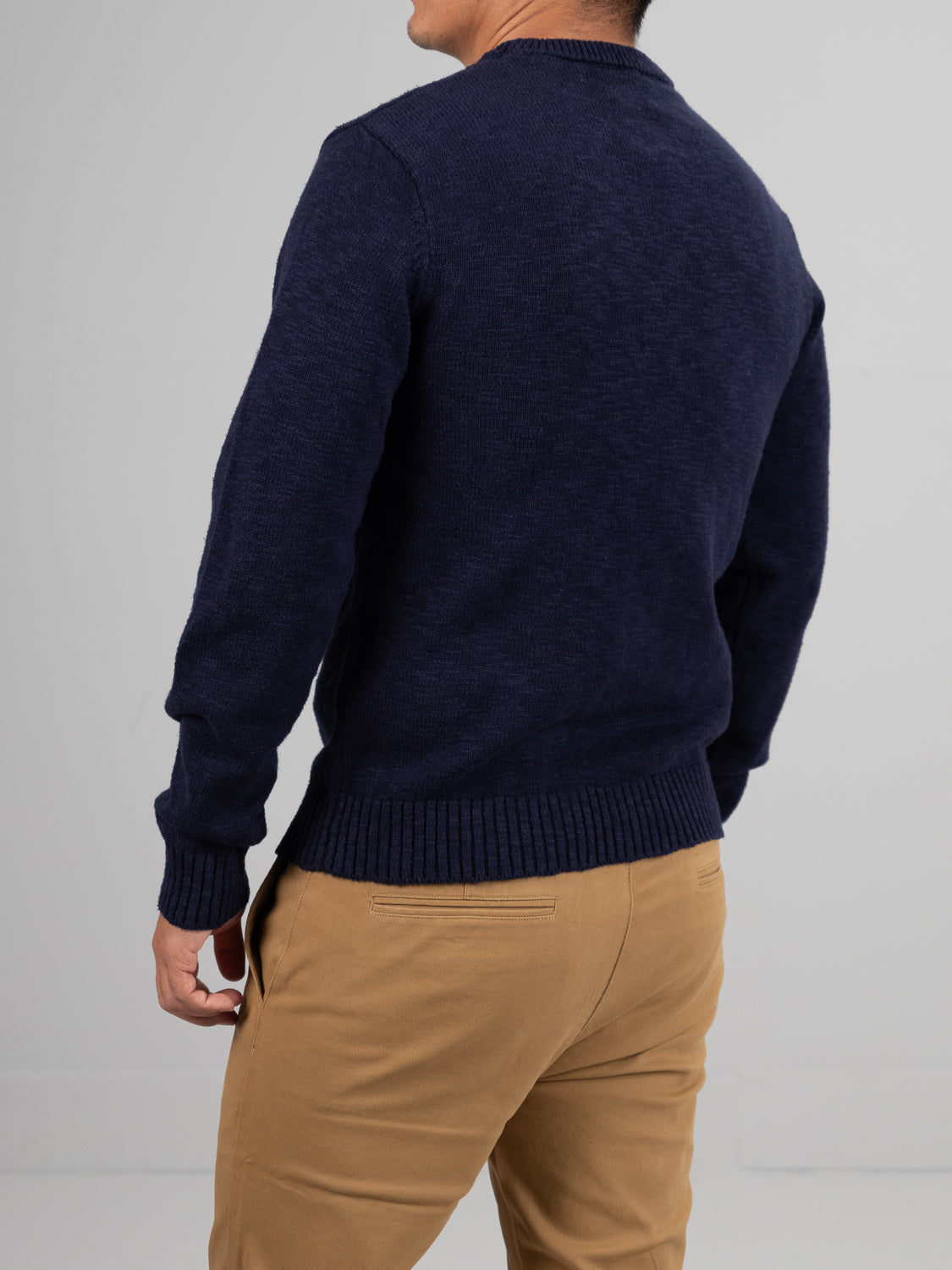 A person in ID Menswears Odense Slub Yarn Sweater, navy blue crew neck, regular fit, and tan pants stands with their back to the camera against a plain gray background.