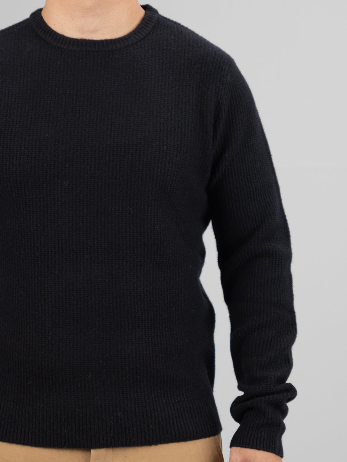 Dressed in ID Menswears Uppsala Merino Wool Sweater, featuring a ribbed popcorn knit stitch and round neckline, paired with tan pants, the person stands against a light gray backdrop. The cozy black sweater is perfect for chilly weather, but their face remains unseen.