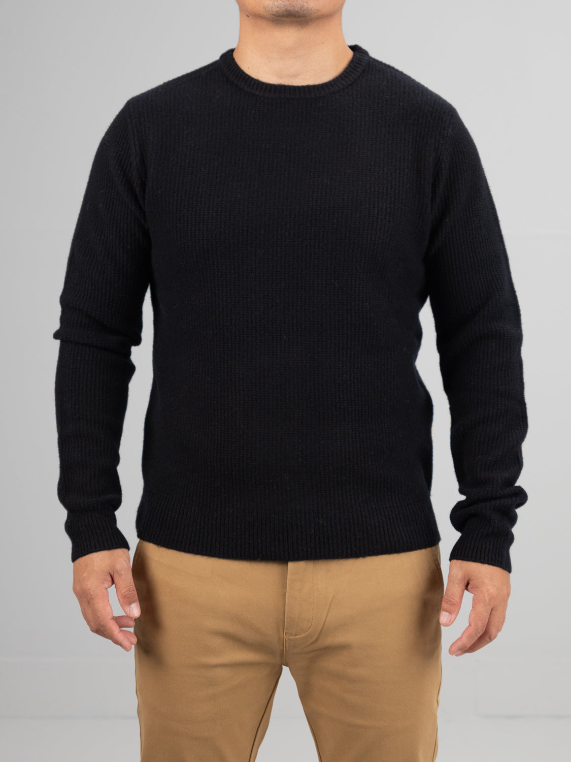 A person in a black Uppsala Merino Wool Sweater by ID Menswear and tan pants stands against a gray backdrop. The ribbed popcorn knit texture is perfect for cold days, keeping the focus on the clothing as their face is not shown.