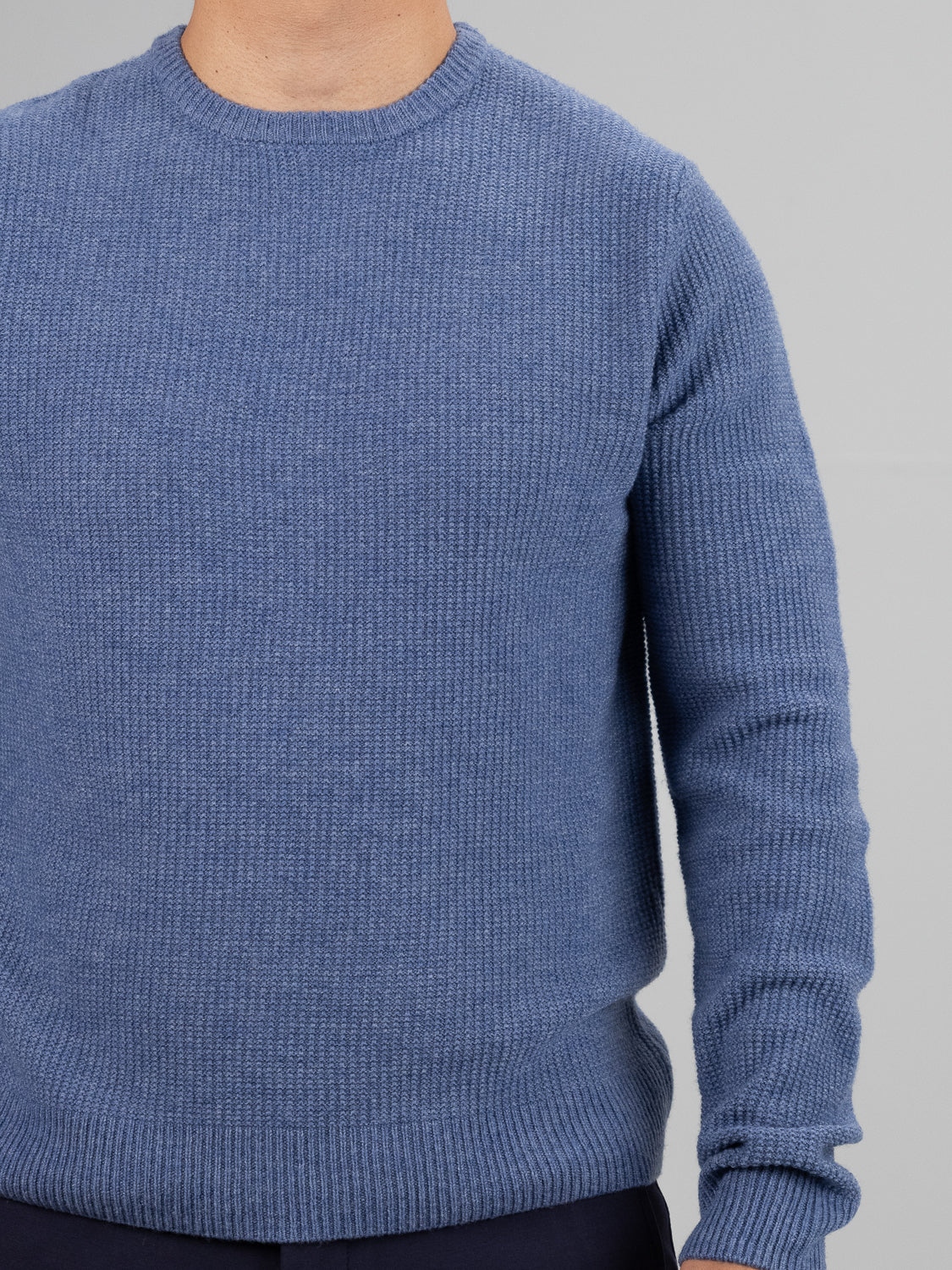Someone is wearing the Uppsala Merino Wool Sweater by ID Menswear, featuring a ribbed popcorn knit stitch in blue. The photo captures only the upper body and part of the arms against a neutral gray background, highlighting this chilly weather attire.