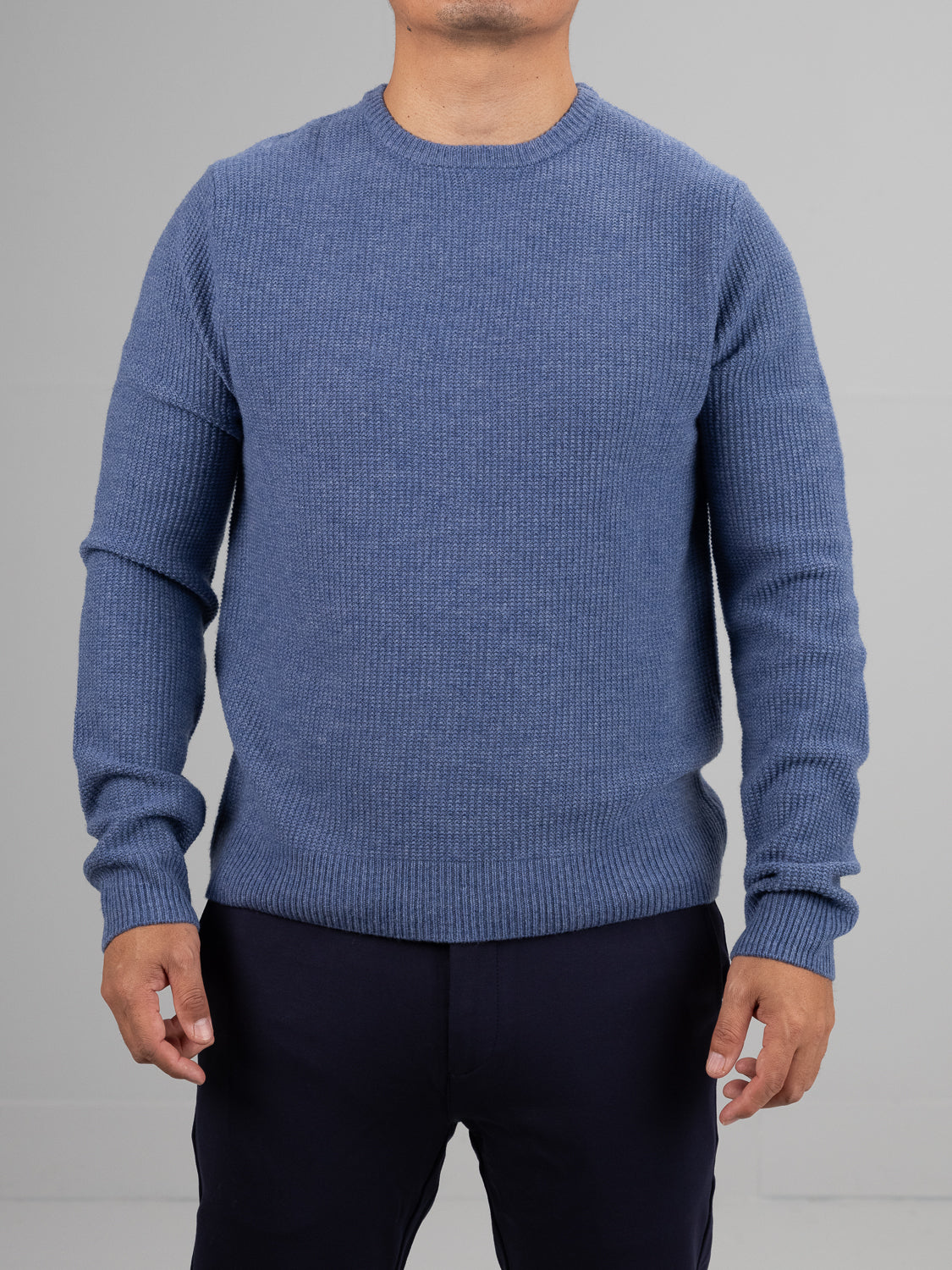 Wearing the blue Uppsala Merino Wool Sweater by ID Menswear, a person stands against a gray background in dark pants, hands relaxed at their sides. Their face isnt visible, but the cozy sweater signals readiness for chilly weather.