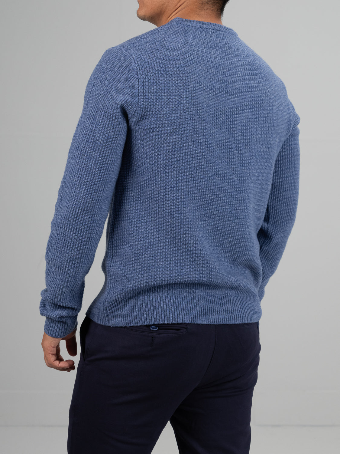 Someone in an ID Menswear Uppsala Merino Wool Sweater and dark pants stands back to the camera against a plain, light gray background. Their right hand is partially visible at their side, prepared for chilly weather.