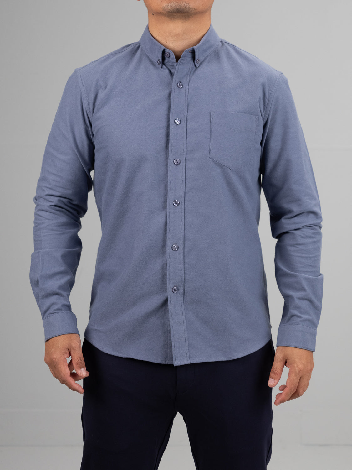 A person is wearing ID Menswears Milo Brushed Oxford Flannel Cotton Shirt, with long sleeves, button-up closure, button-down collar, and a chest pocket. It pairs with dark pants against a plain gray backdrop.