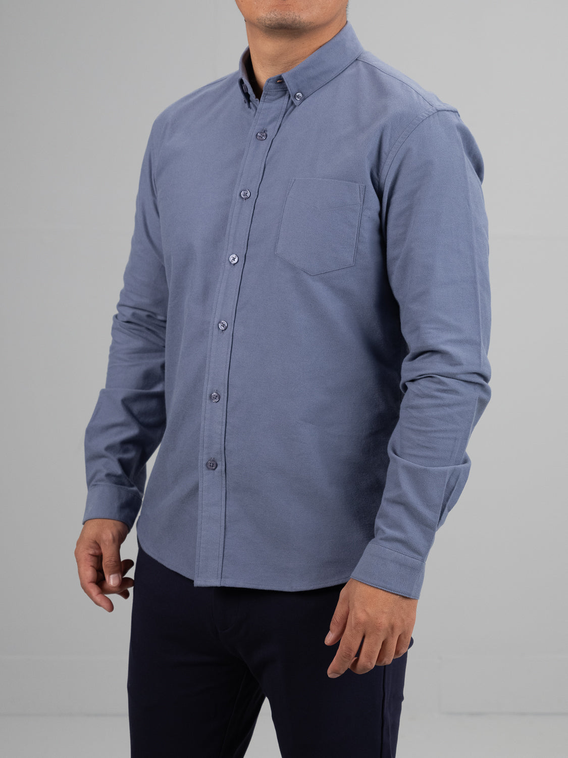 A model wears the Milo Brushed Oxford Flannel Cotton Shirt by ID Menswear, featuring long sleeves and a left-side pocket in light gray, paired with dark pants against a plain gray background.