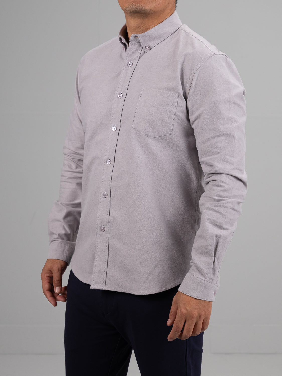 A model poses against a plain gray backdrop, wearing ID Menswears Milo Brushed Oxford Flannel Cotton Shirt in light gray. The shirt features a front pocket and neatly fastened long sleeves, paired with dark pants. The models face is not visible.