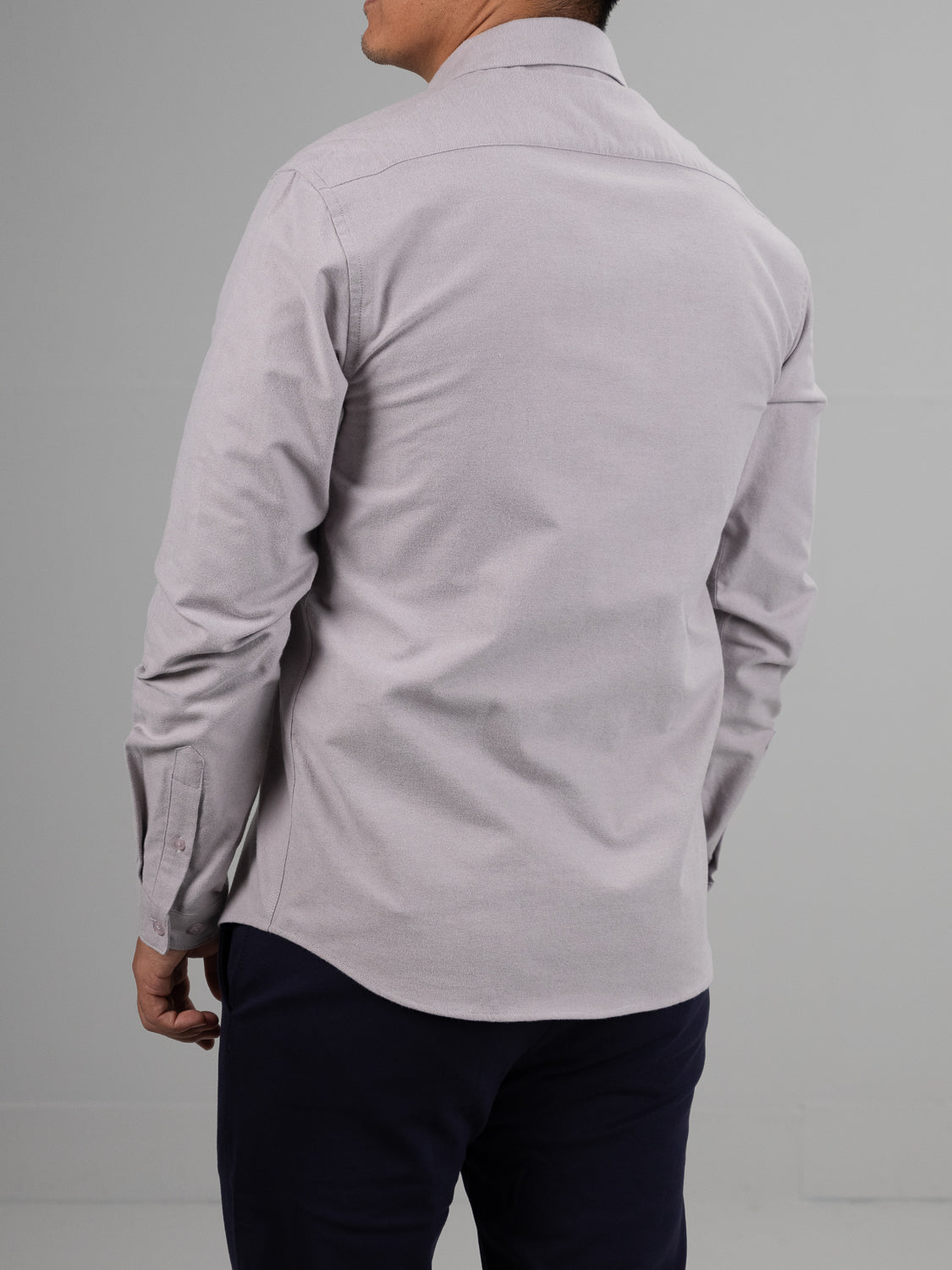A person wearing the ID Menswear Milo Brushed Oxford Flannel Cotton Shirt in light gray, with long sleeves, and dark pants is seen from the back against a plain, neutral background.