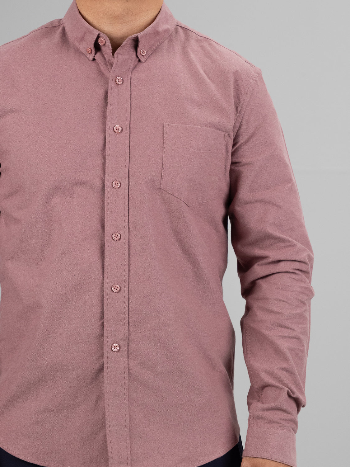 A person models the Milo Brushed Oxford Flannel Cotton Shirt by ID Menswear, featuring a long-sleeved, button-up mauve design with a front pocket against a plain gray background. The versatile cotton shirt is tucked into trousers, and the head is not visible in the image.