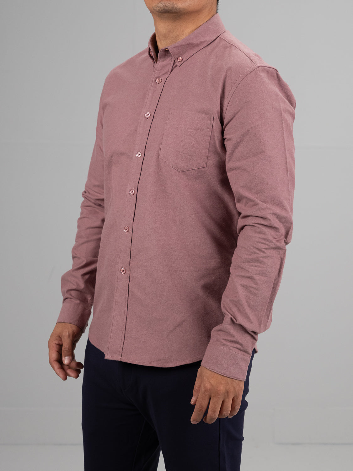 Against a light gray background, a person in a Milo Brushed Oxford Flannel Cotton Shirt by ID Menswear, featuring a dusty pink hue with long sleeves and front pocket, is paired with dark pants.