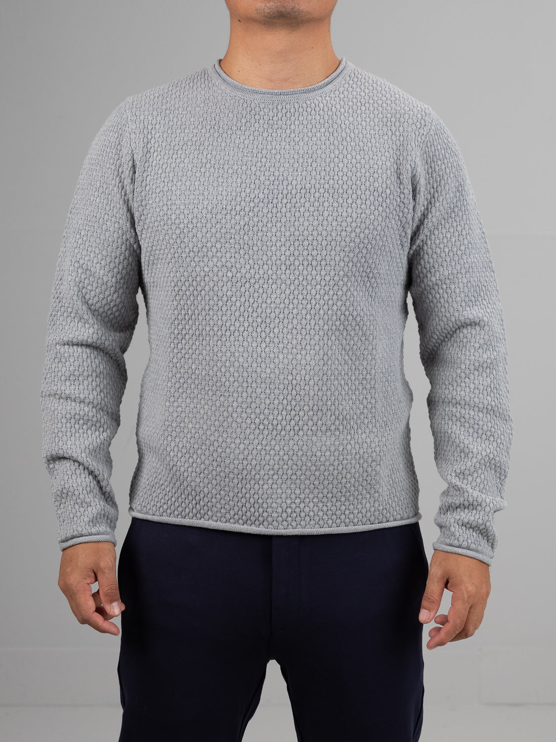 Against a plain gray backdrop, a person wears an ID Menswears Westside Rolled Neck Sweater in light gray with a subtle pattern and dark pants. Their hands rest relaxed at their sides, though their head is not visible. Ideal for casual knitwear lovers.
