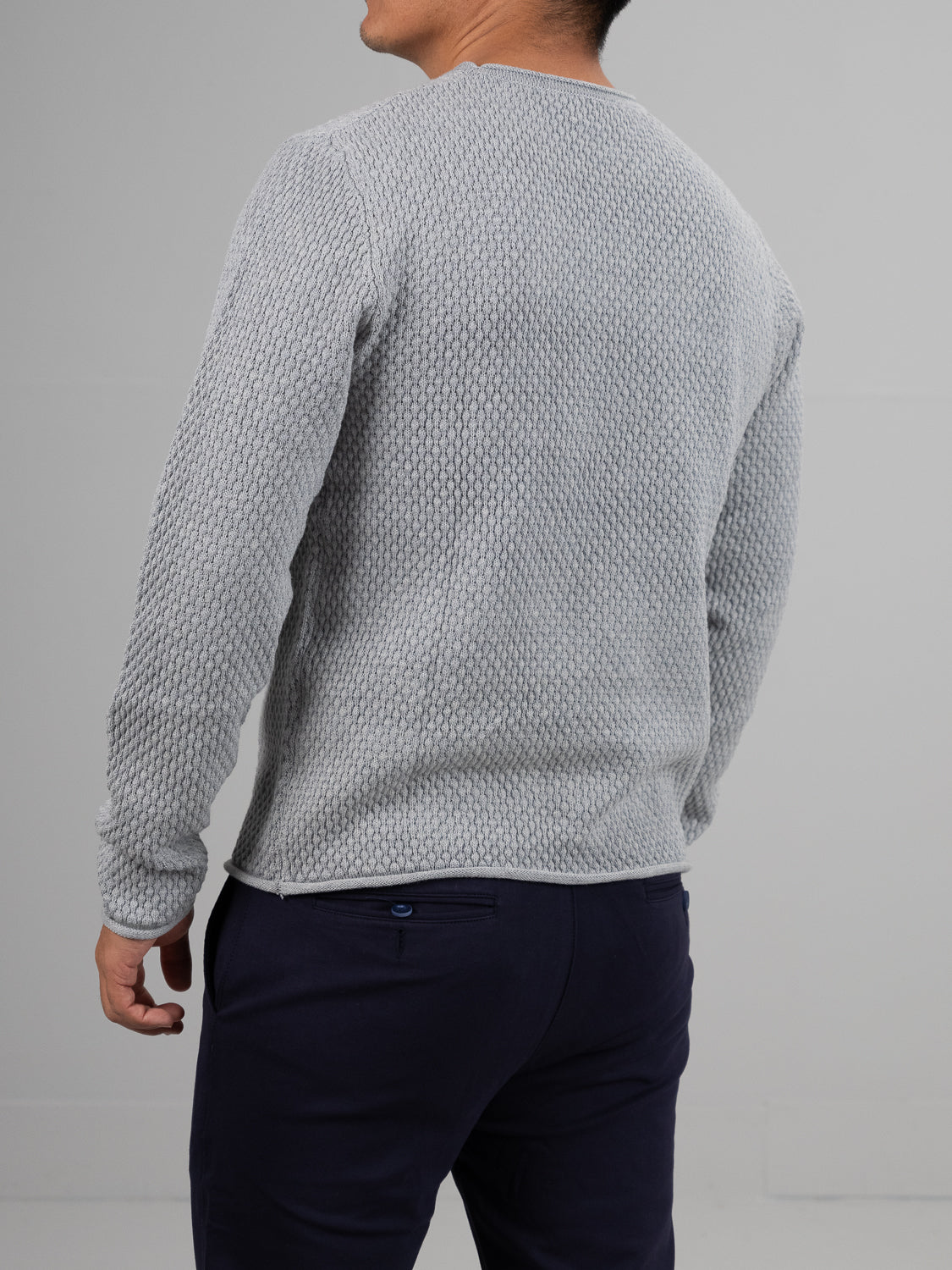 Someone in a Westside Rolled Neck Sweater from ID Menswear, textured gray, paired with dark pants, faces away against a plain light gray wall, epitomizing casual knitwear at its best.