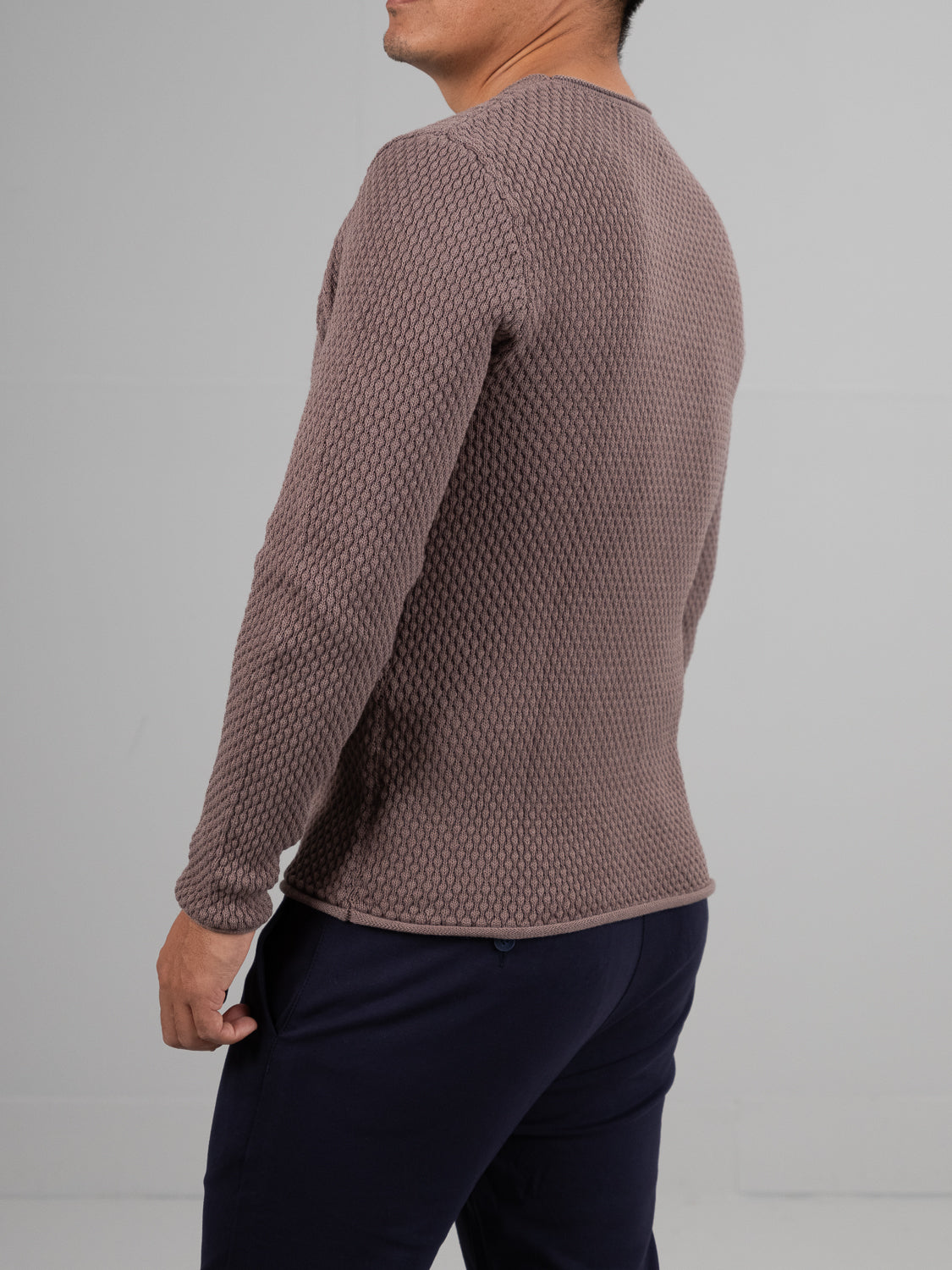 A person wearing a textured brown Westside Rolled Neck Sweater by ID Menswear and dark pants stands against a plain gray background, slightly turned to the side with their right arm partially in view.