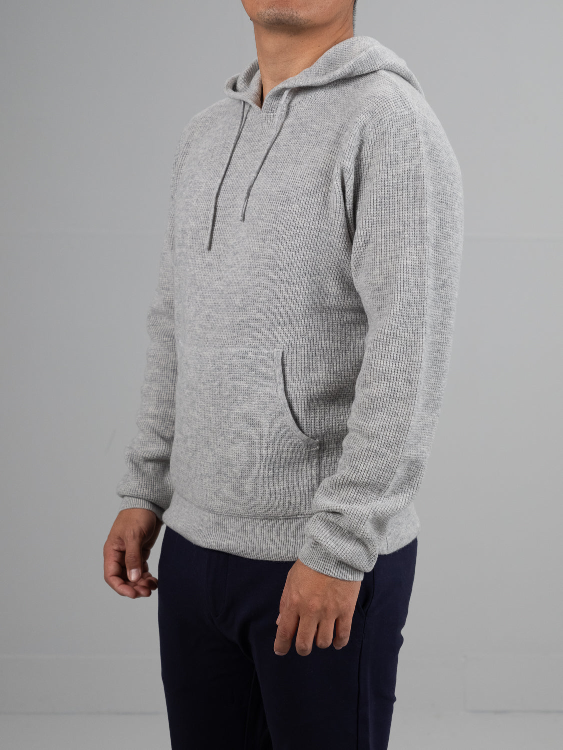 Someone in a light gray Bergen Waffle Knit Merino Hoody from ID Menswear stands against a plain background. The hoody features a front pocket and drawstrings, paired with dark pants. Their face is not visible.