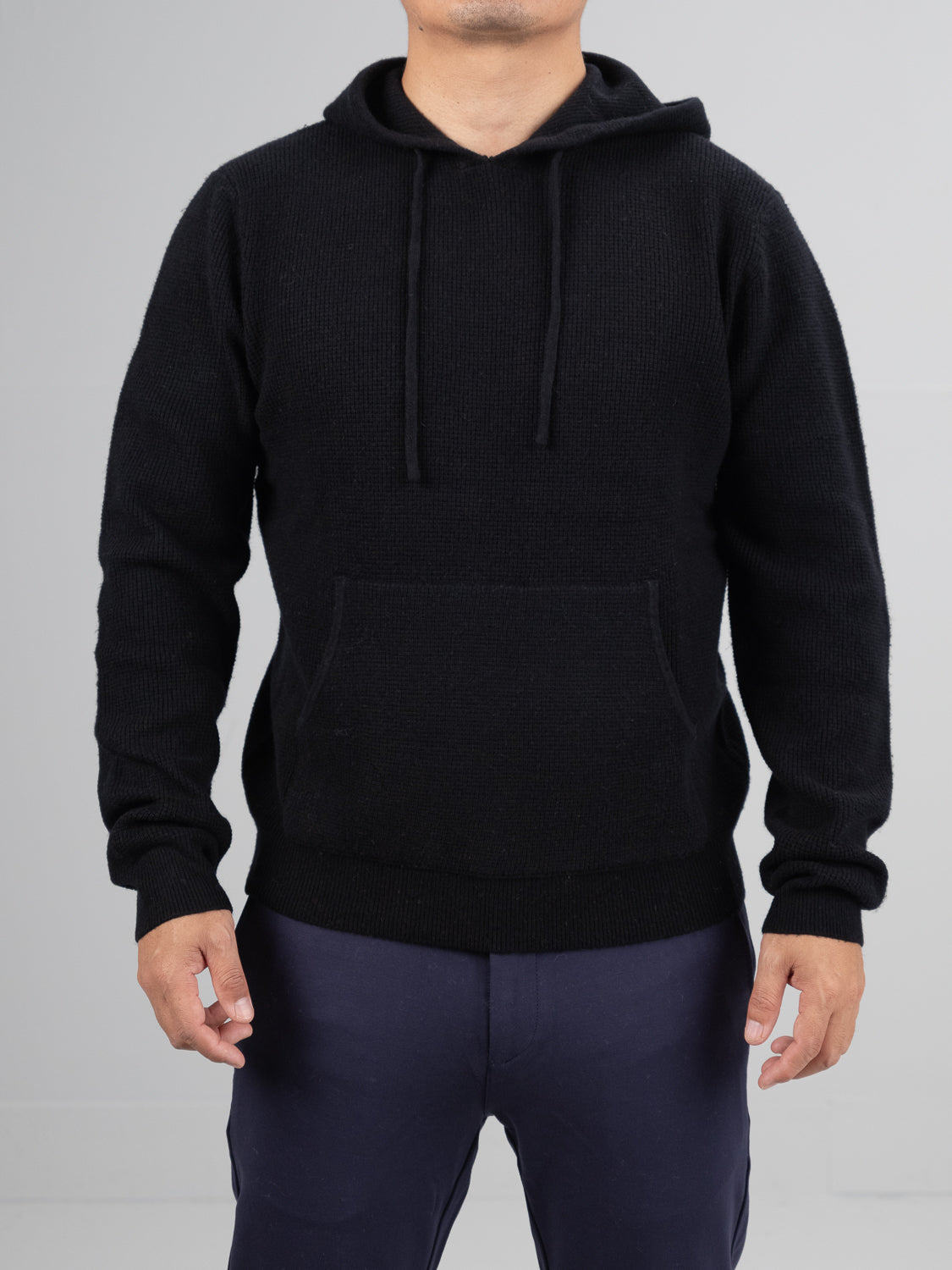 Someone wears the Bergen Waffle Knit Merino Hoody by ID Menswear with navy pants, standing against a plain gray background. The black hoody has a front pocket and drawstrings. Their hands are relaxed by their sides, and the face is not visible.
