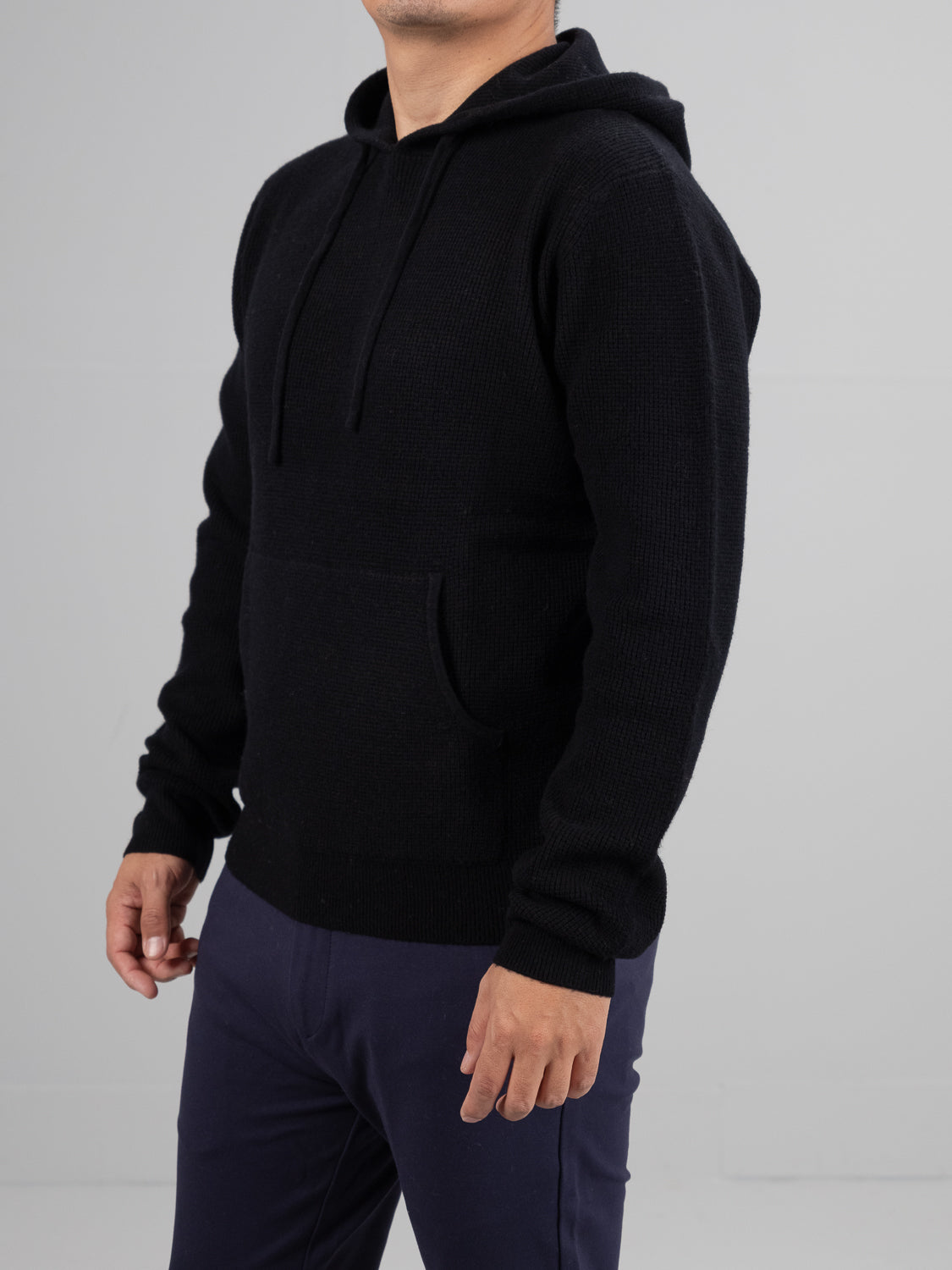 A person wears an ID Menswear Bergen Waffle Knit Merino Hoody in black, featuring a front pocket and drawstrings, standing against a plain gray background with their left hand partially visible by their side. They pair it with dark pants.